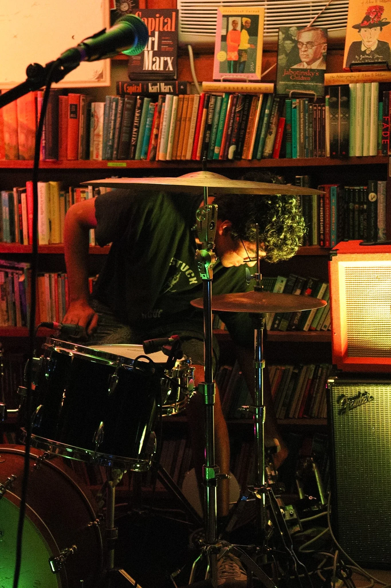 A person with curly hair and glasses, wearing a dark shirt, is playing drums in a room filled with books and musical equipment. There is a microphone and a drum set in the foreground, and the background features a bookshelf with various books and pos