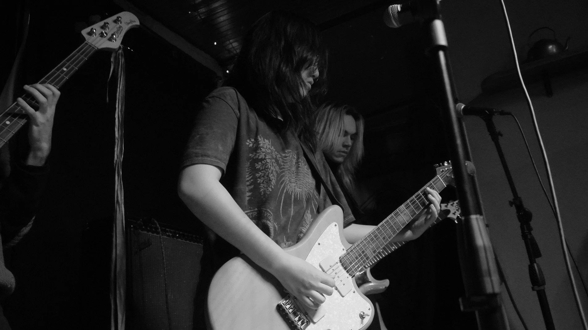 Black and white photo of two female musicians playing electric guitars on stage. They are focused on their instruments, with microphones nearby.