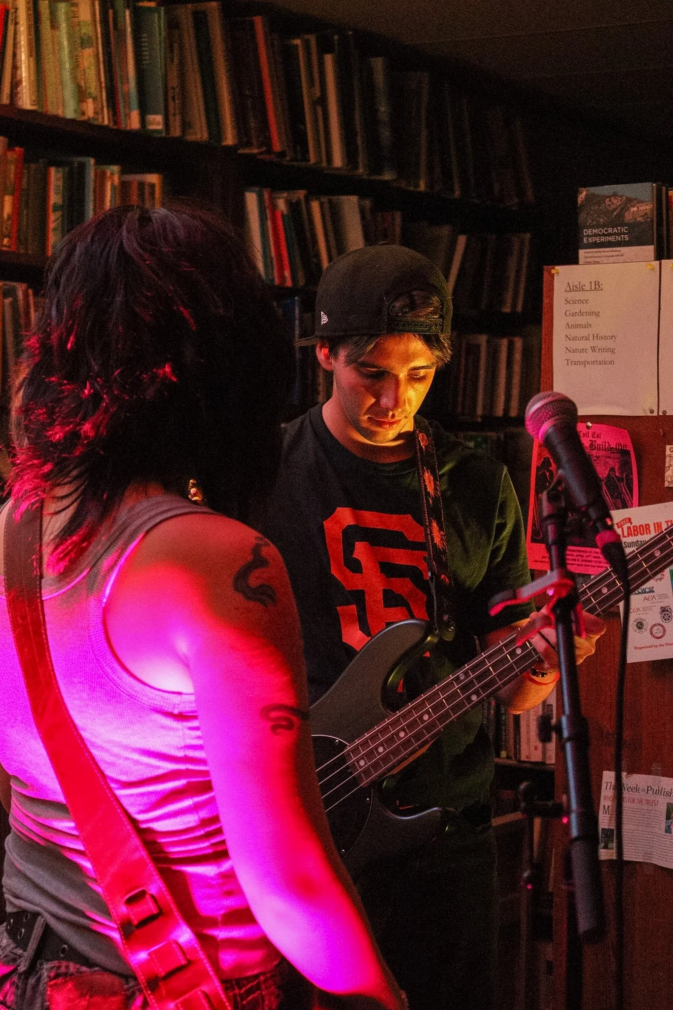 A young man is playing an electric guitar indoors, standing near a woman with tattoos on her arm, in front of a bookshelf and posters on the wall.