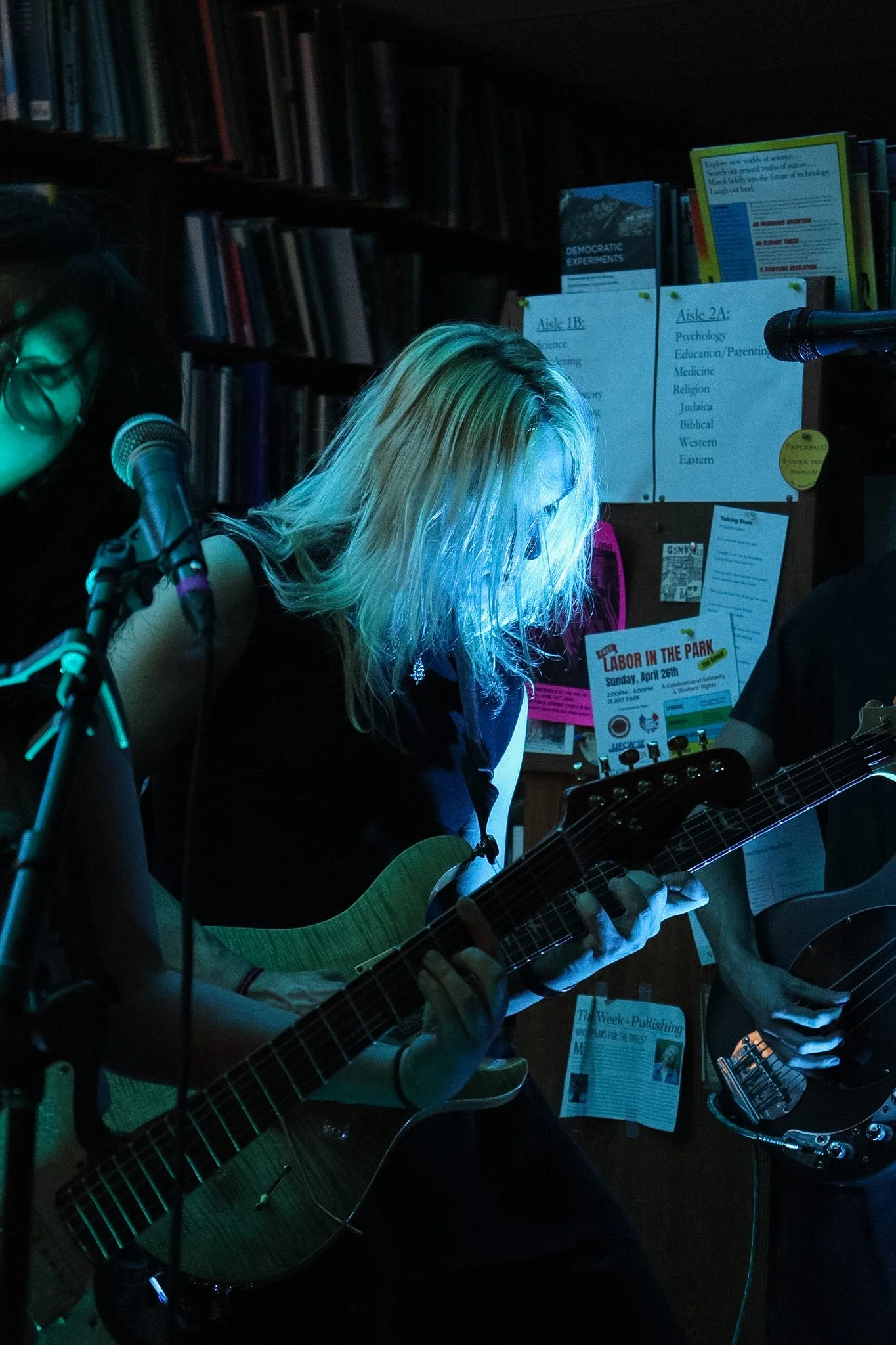 A person with blonde hair playing a guitar at a music event with a woman singing into a microphone nearby. The background includes bookshelves and promotional posters.