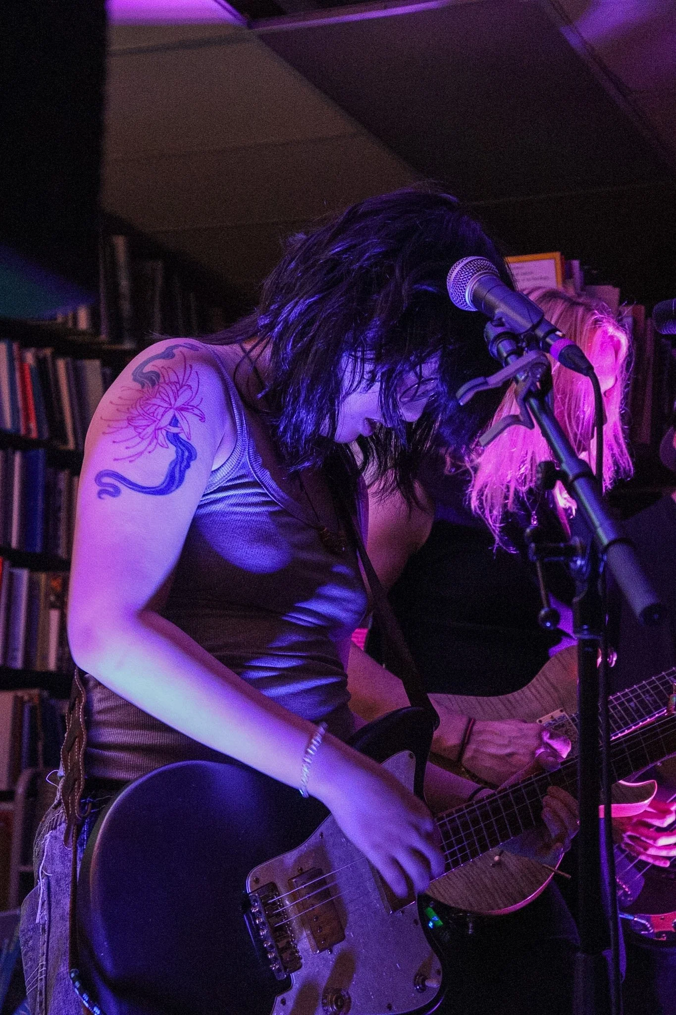 Two musicians performing in a dimly lit, purple-hued room with bookshelves in the background. One is playing an electric guitar, while the other appears to be singing into a microphone.
