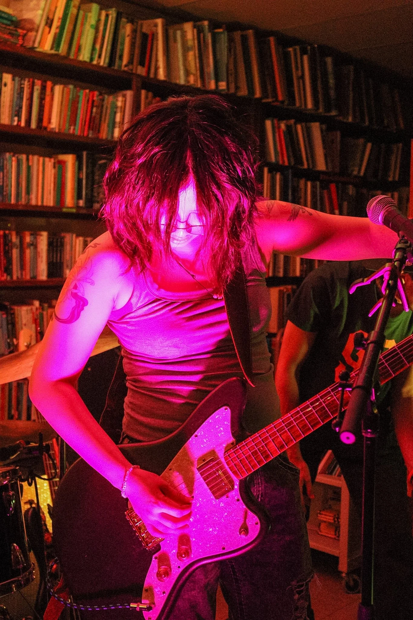 A person with curly hair playing an electric guitar in a dimly lit room with bookshelves in the background, illuminated by pink stage lighting.