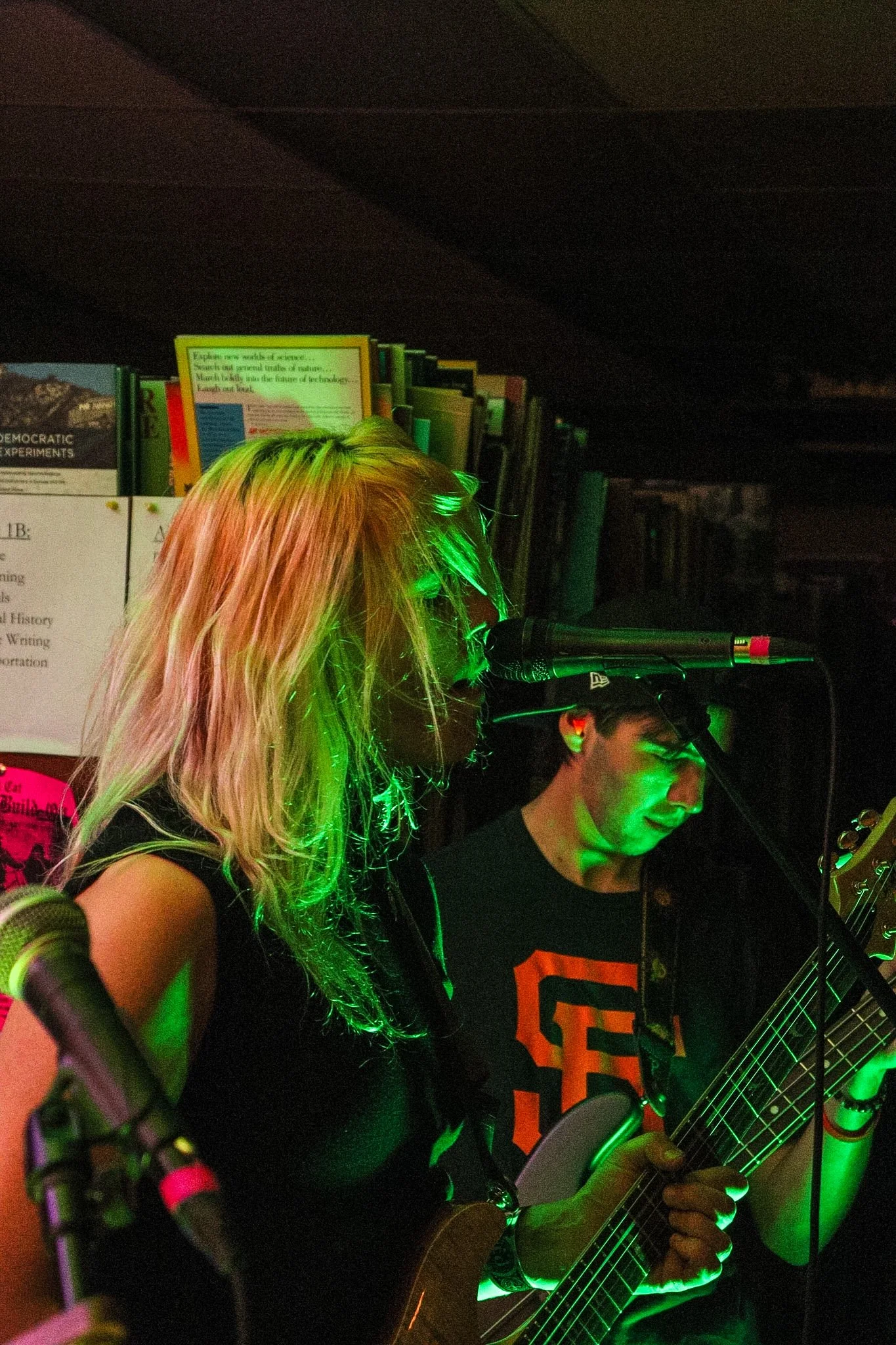 Two musicians playing guitars in a dimly lit room with green and pink lighting. One woman with long, blonde hair and a black shirt is in the foreground, and a man with glasses and a black shirt with orange lettering is in the background, also playing