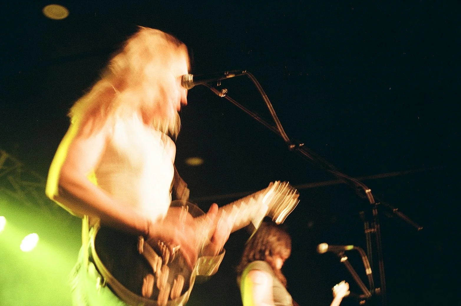 A musician with long hair playing an electric guitar on stage, with lighting and microphone stands visible.