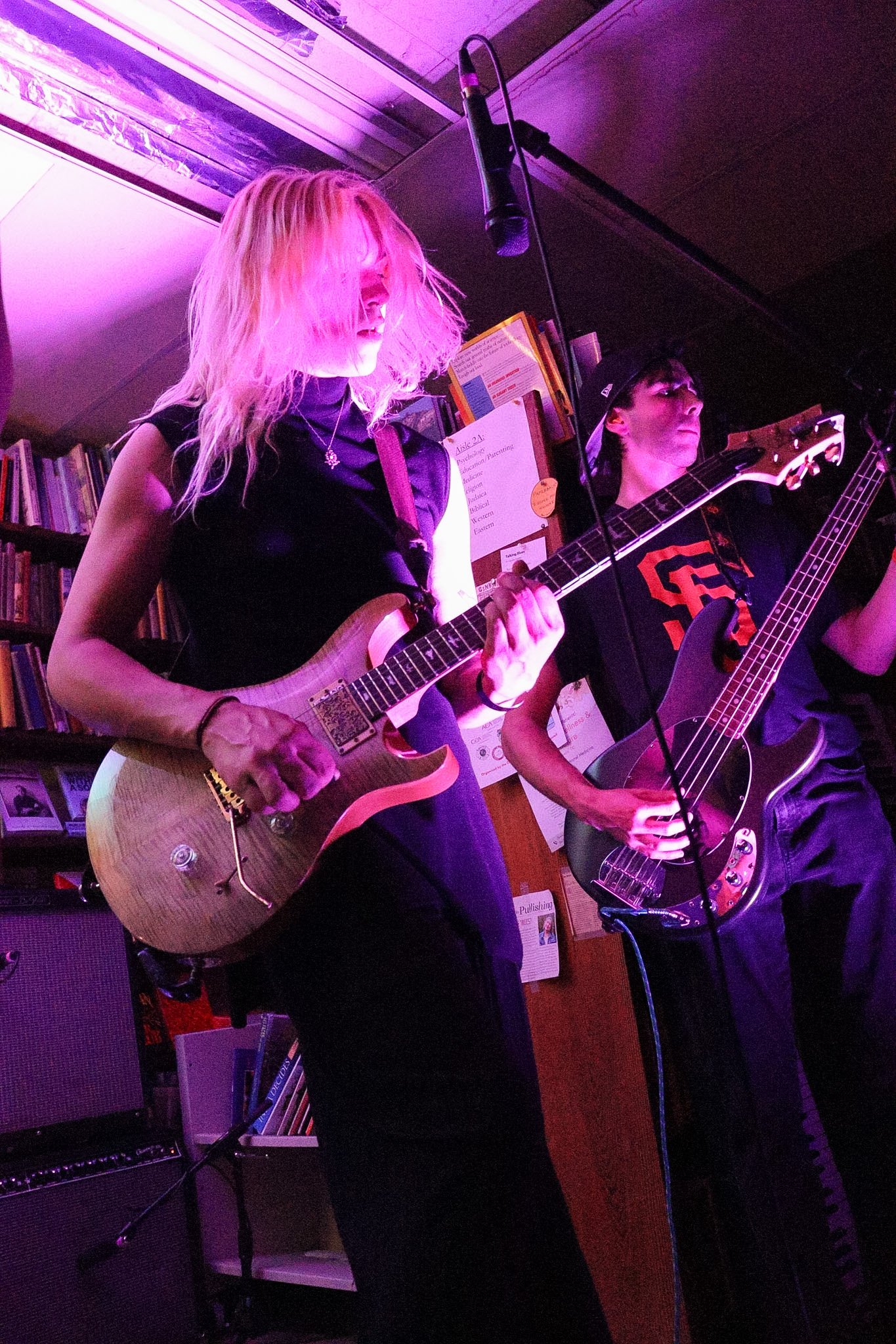 Two musicians performing with electric guitars in a cozy, bookshelf-lined room with purple and pink lighting. One female guitarist with blond hair and black clothing, and one male guitarist wearing a black cap and shirt.