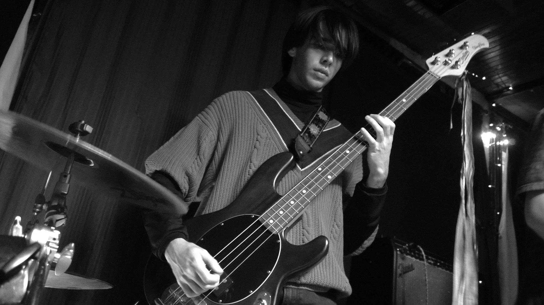 A person with medium-length dark hair playing a bass guitar in a dimly lit room, possibly a music studio or performance space.