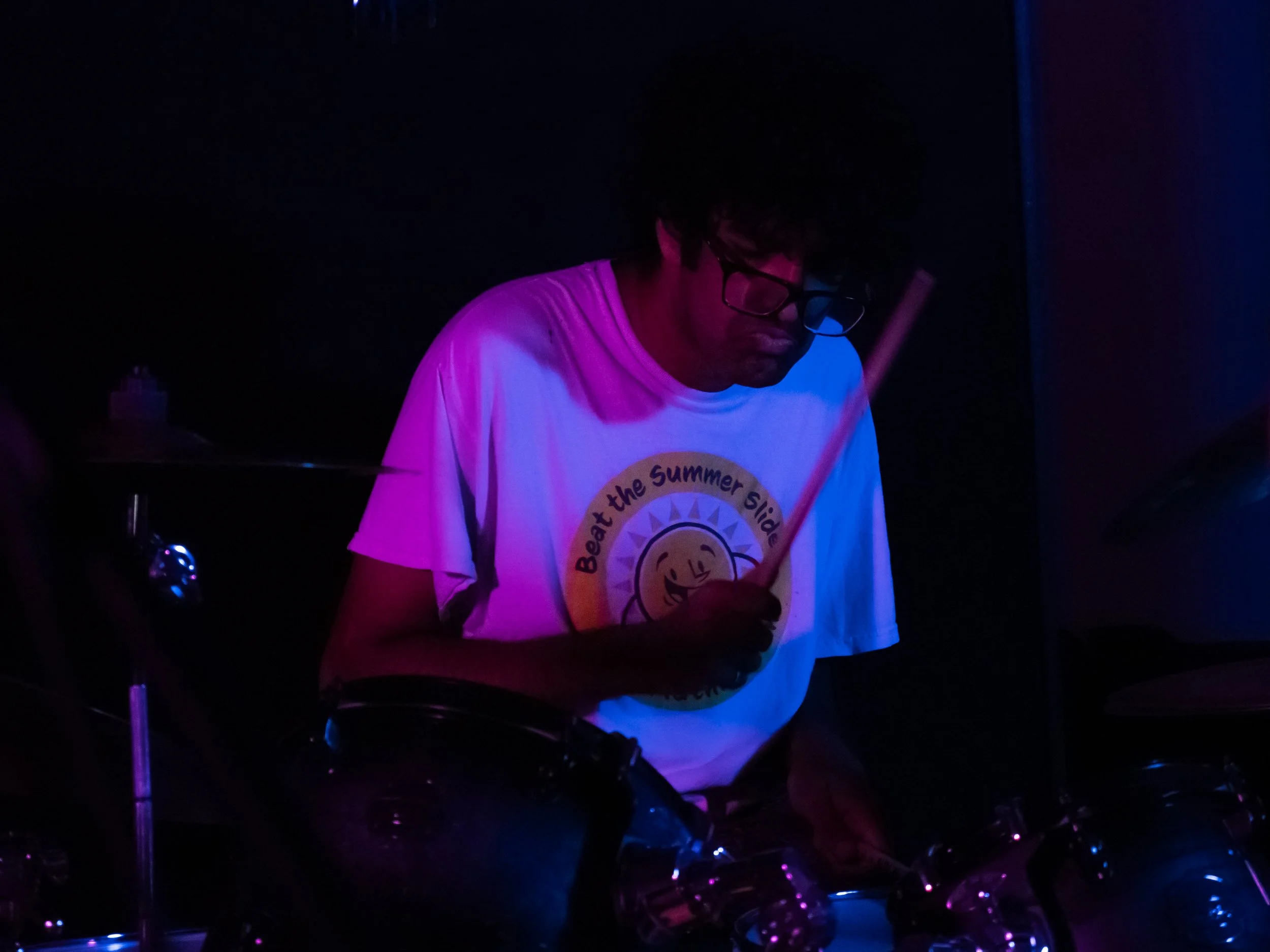 Person playing drums in a dark room illuminated by purple and blue lights, wearing glasses and a t-shirt with a smiling sun logo and the text 'Beat the Summer Slide'.