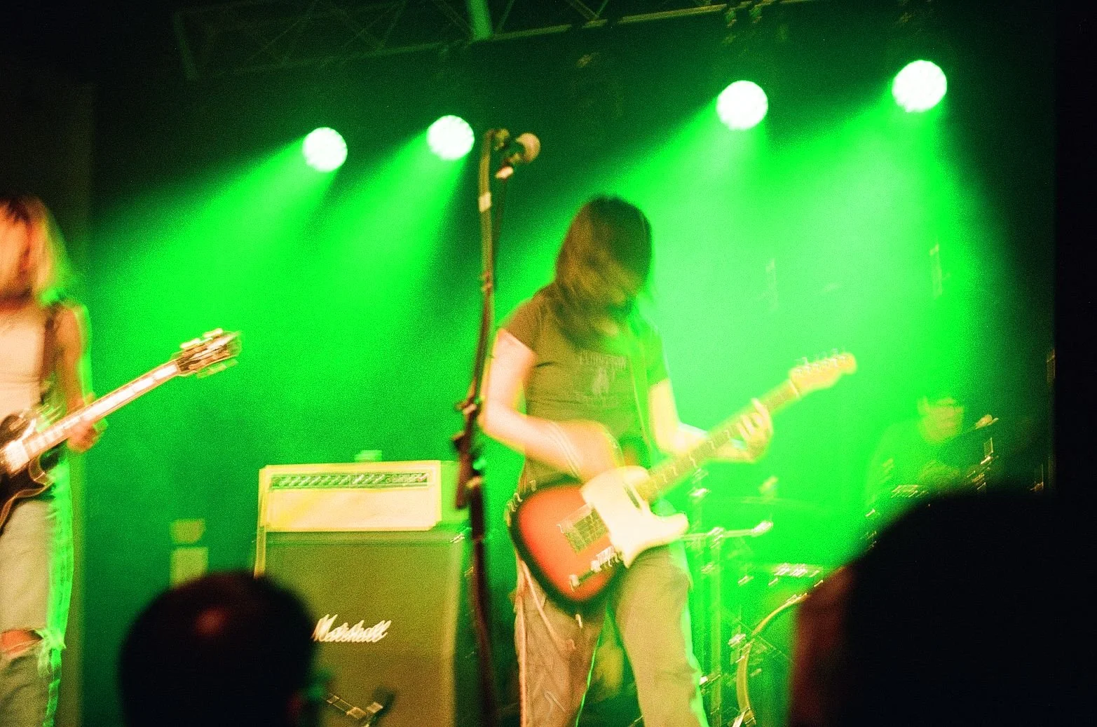 A band performs on stage with green stage lighting, featuring two guitarists in the foreground and a drummer in the background.