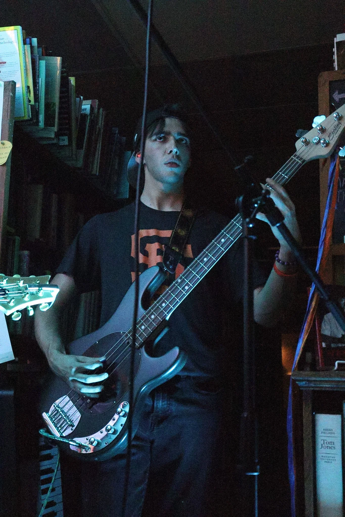 A person playing an electric bass guitar in a dimly lit room, surrounded by books and musical equipment.
