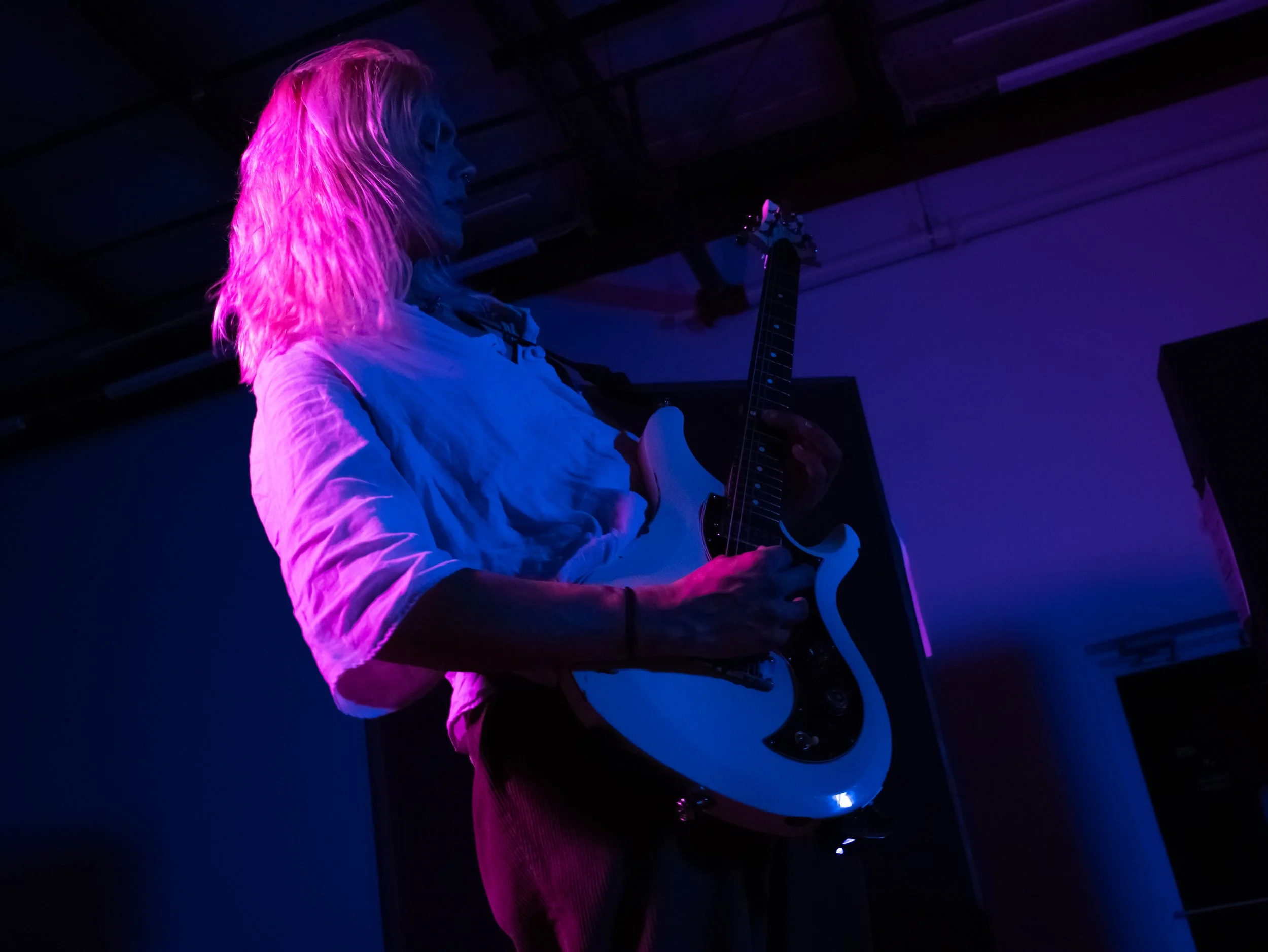 Person with long blonde hair playing an electric guitar on stage, illuminated by pink and purple stage lights.