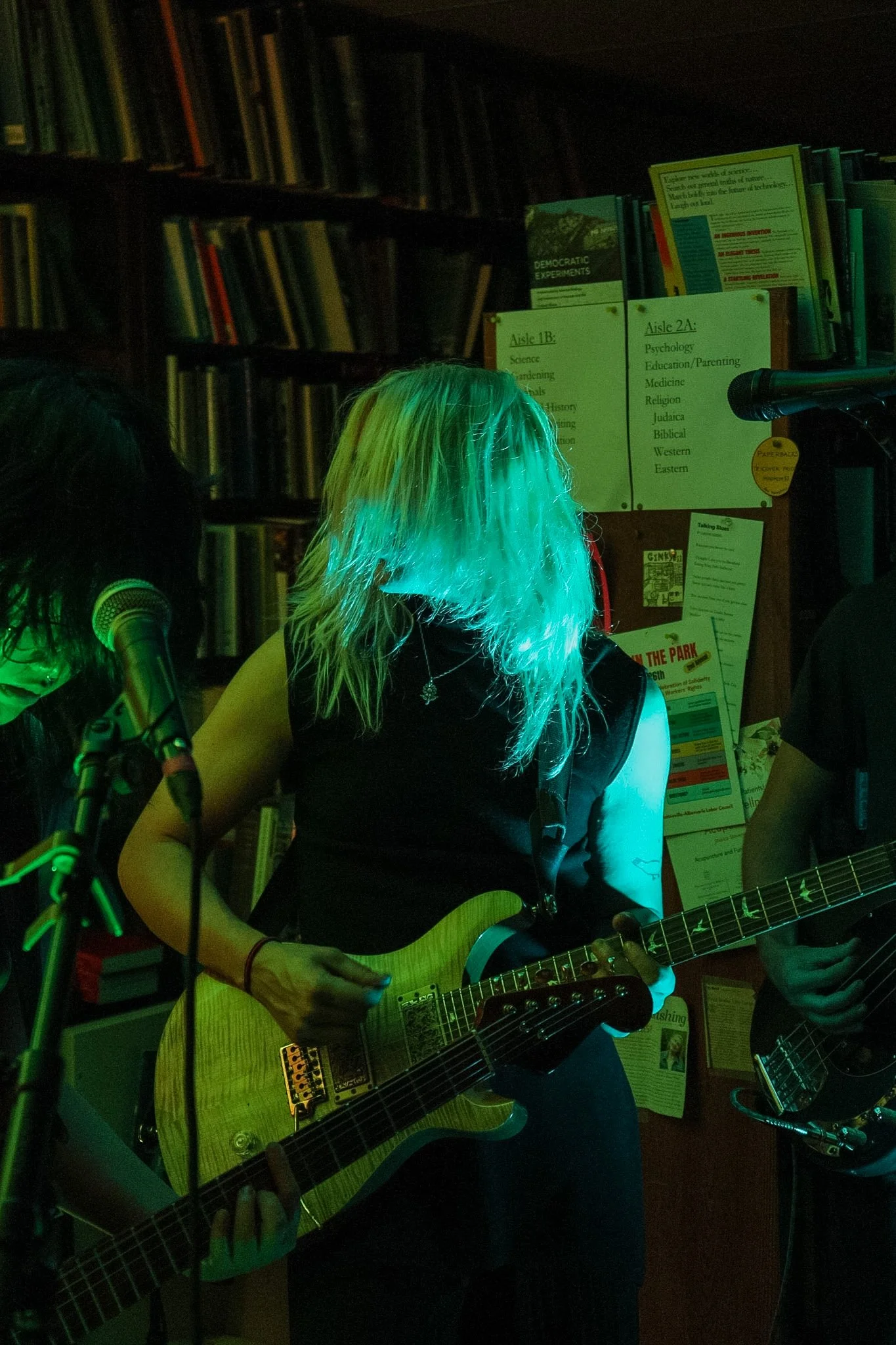 Person with dyed green hair playing an electric guitar at a music venue with a bookshelf and musical equipment in the background.