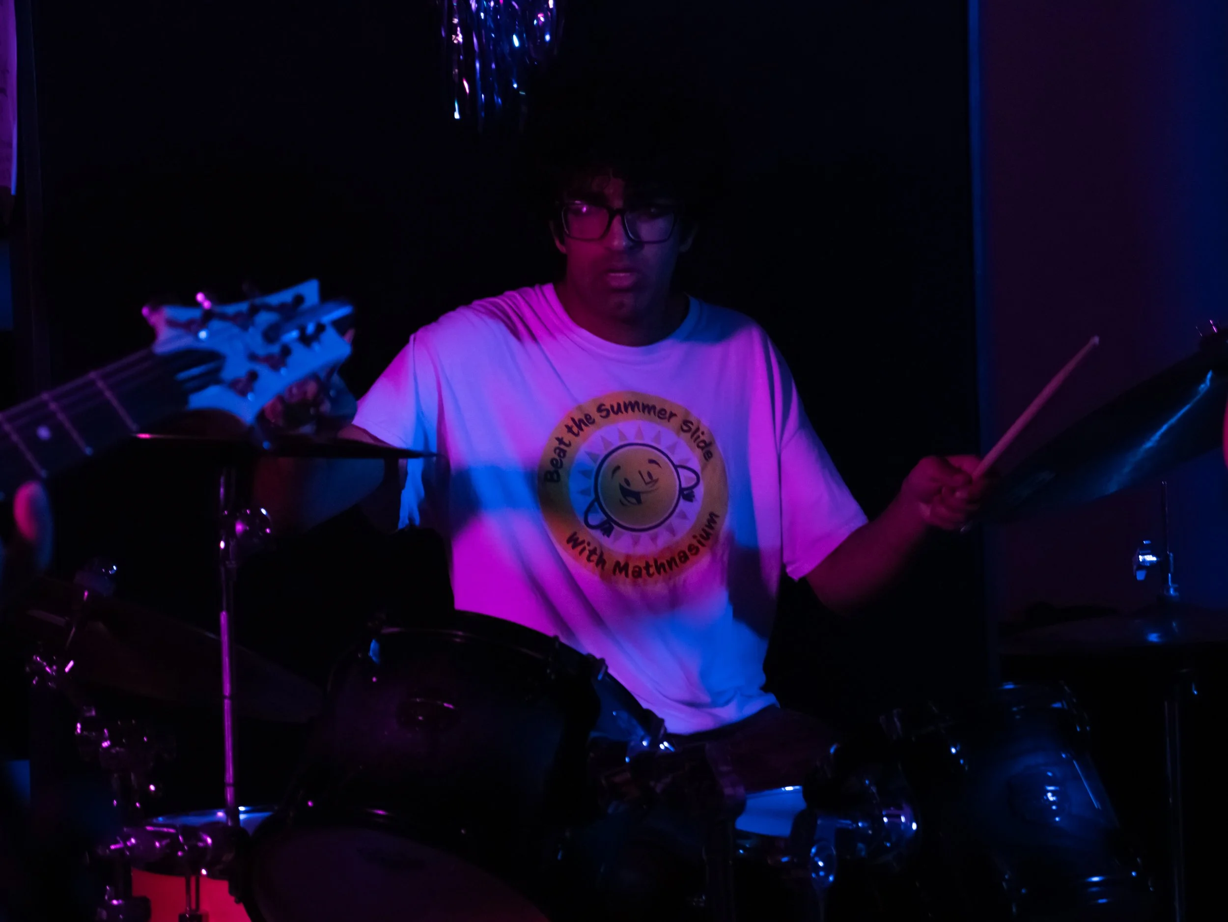 A person with black hair and glasses playing drums in a dark room illuminated by purple and blue lights. The person is wearing a white t-shirt with a yellow smiley face and the text 'Beach the Summer Sides with Mathnasium'.