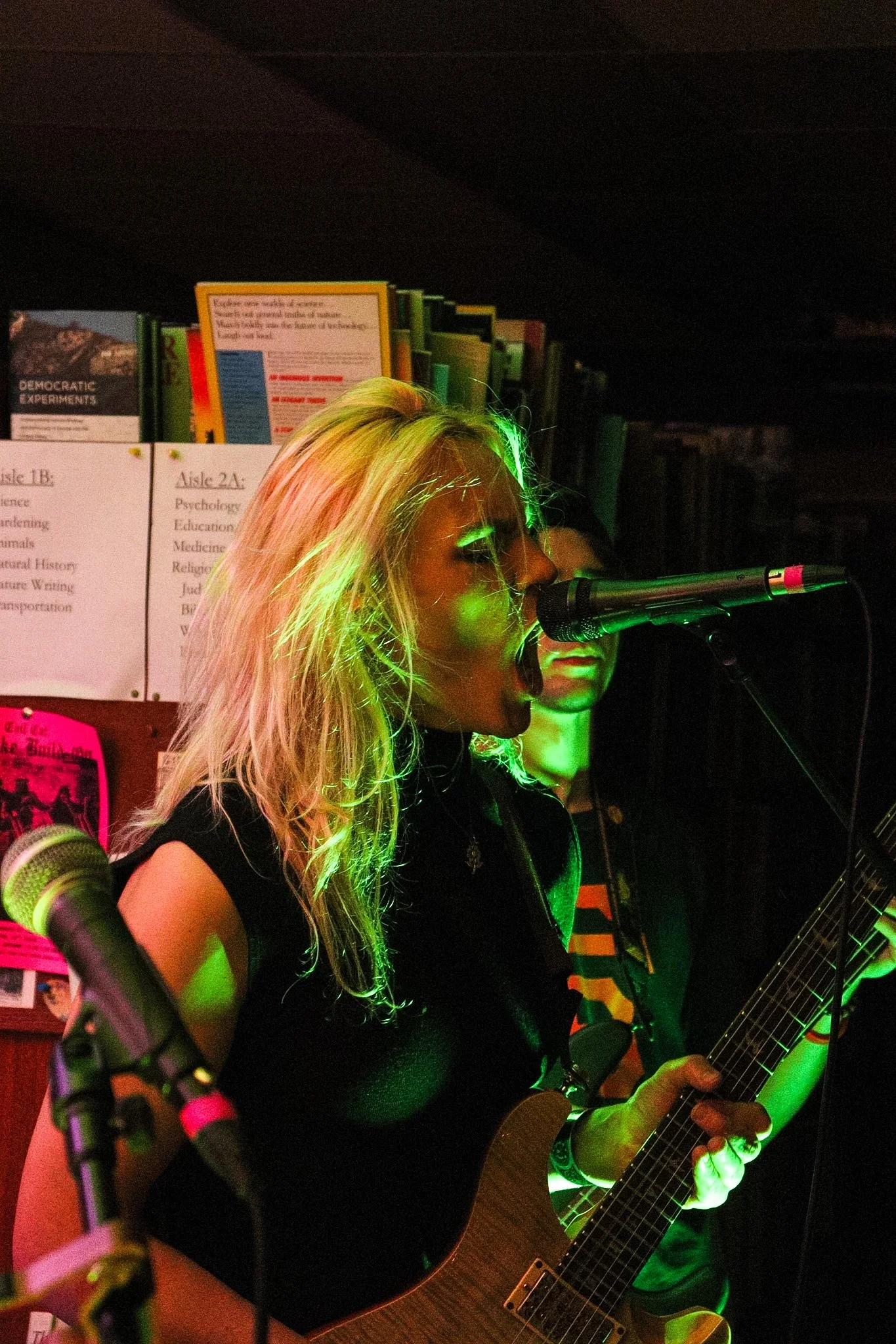 A woman with blonde hair playing an acoustic guitar and singing into a microphone during a live performance in a dark indoor setting, with several posters on the wall behind her.