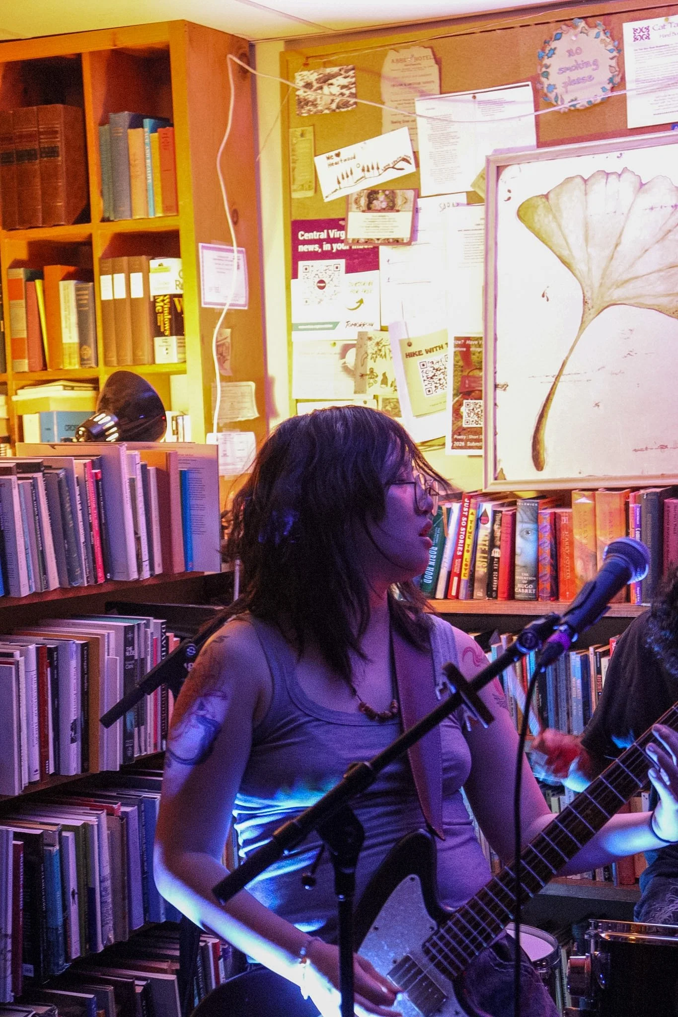 A woman with glasses and long hair playing an electric guitar in a bookstore or library with bookshelves. There are posters and papers on the wall behind her, and a person holding a microphone is partially visible in the foreground.