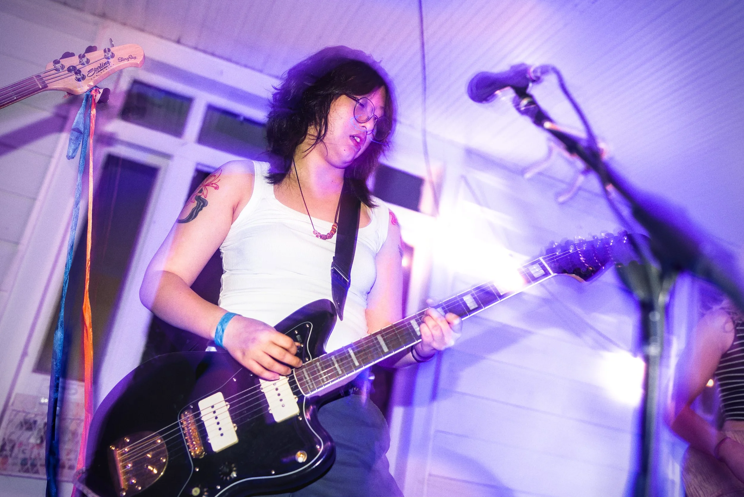 A woman with curly dark hair wearing glasses, a white tank top, and a beaded necklace is playing a black electric guitar with white pickups. She is standing in a room with white paneled walls and purple lighting, near a microphone and a window with m