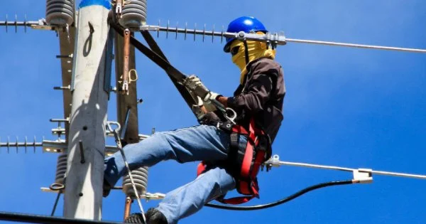 A person in safety gear, including a helmet and harness, working on high-voltage power lines on a utility pole against a clear blue sky.
