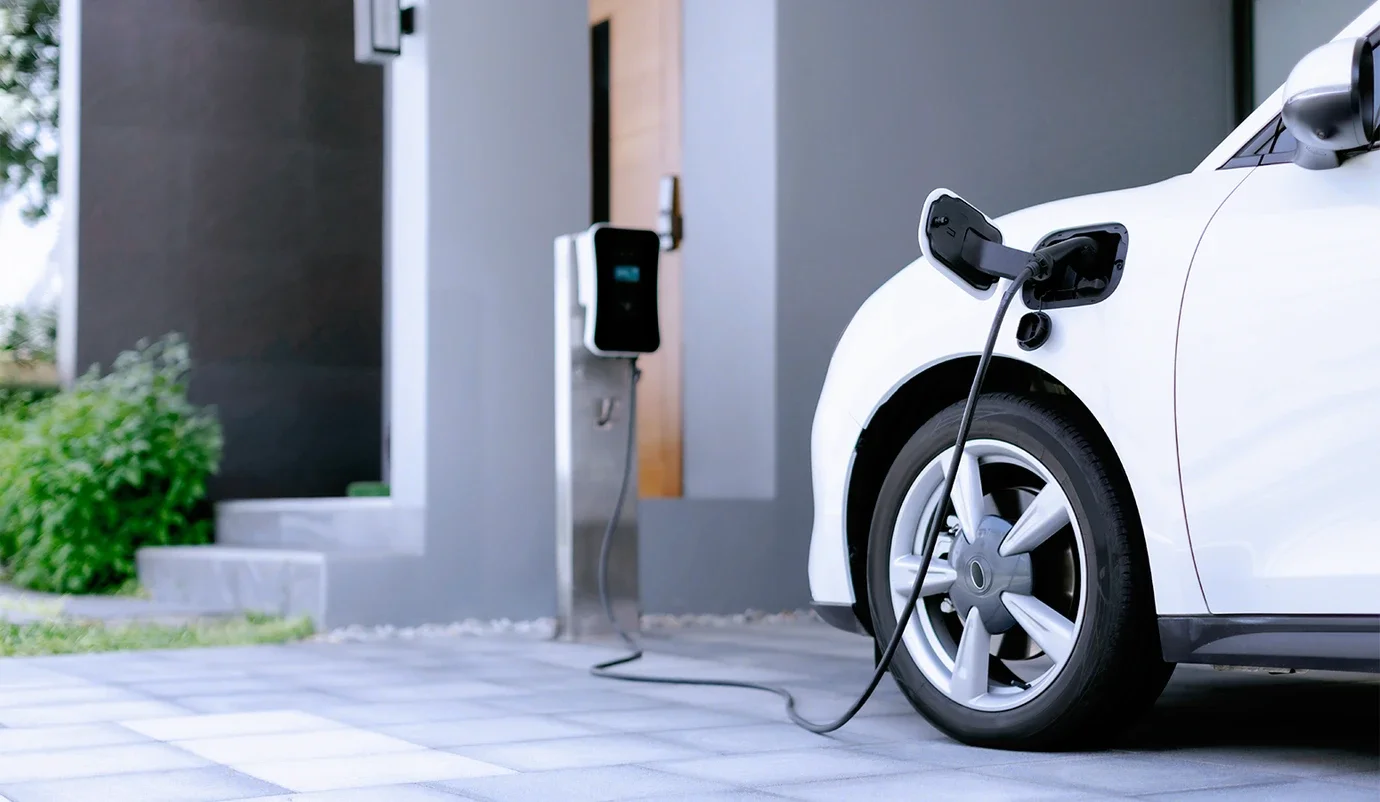 White electric car plugged into a charging station outside a modern building on a paved driveway.