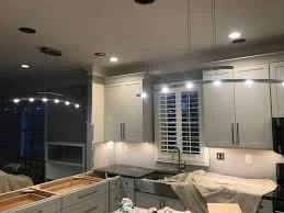 Modern kitchen under construction with white cabinets, a marble countertop, and recessed lighting, with some construction materials and tools visible.