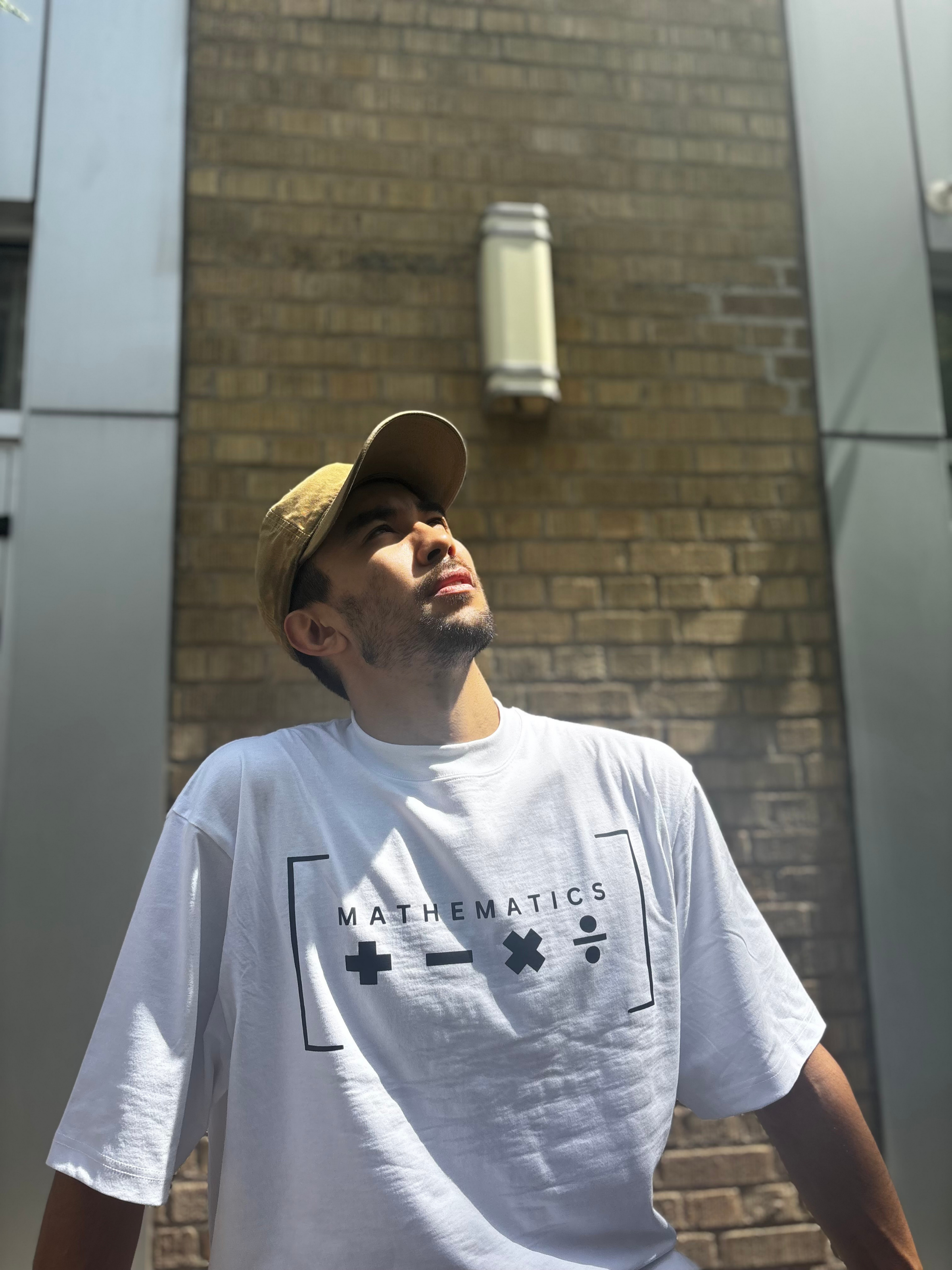 A young man wearing a beige cap and a white T-shirt with black mathematical symbols and the word 'MATHEMATICS' printed on it, standing outdoors in front of a brick building.