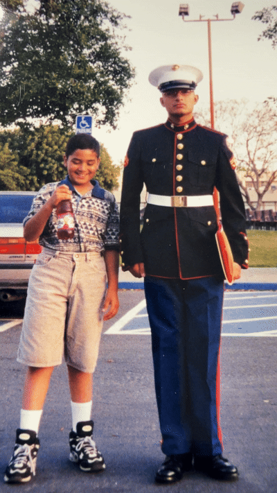 A Hispanic boy holding a bottle of soda standing next to a U.S. Marine in uniform in a parking lot.