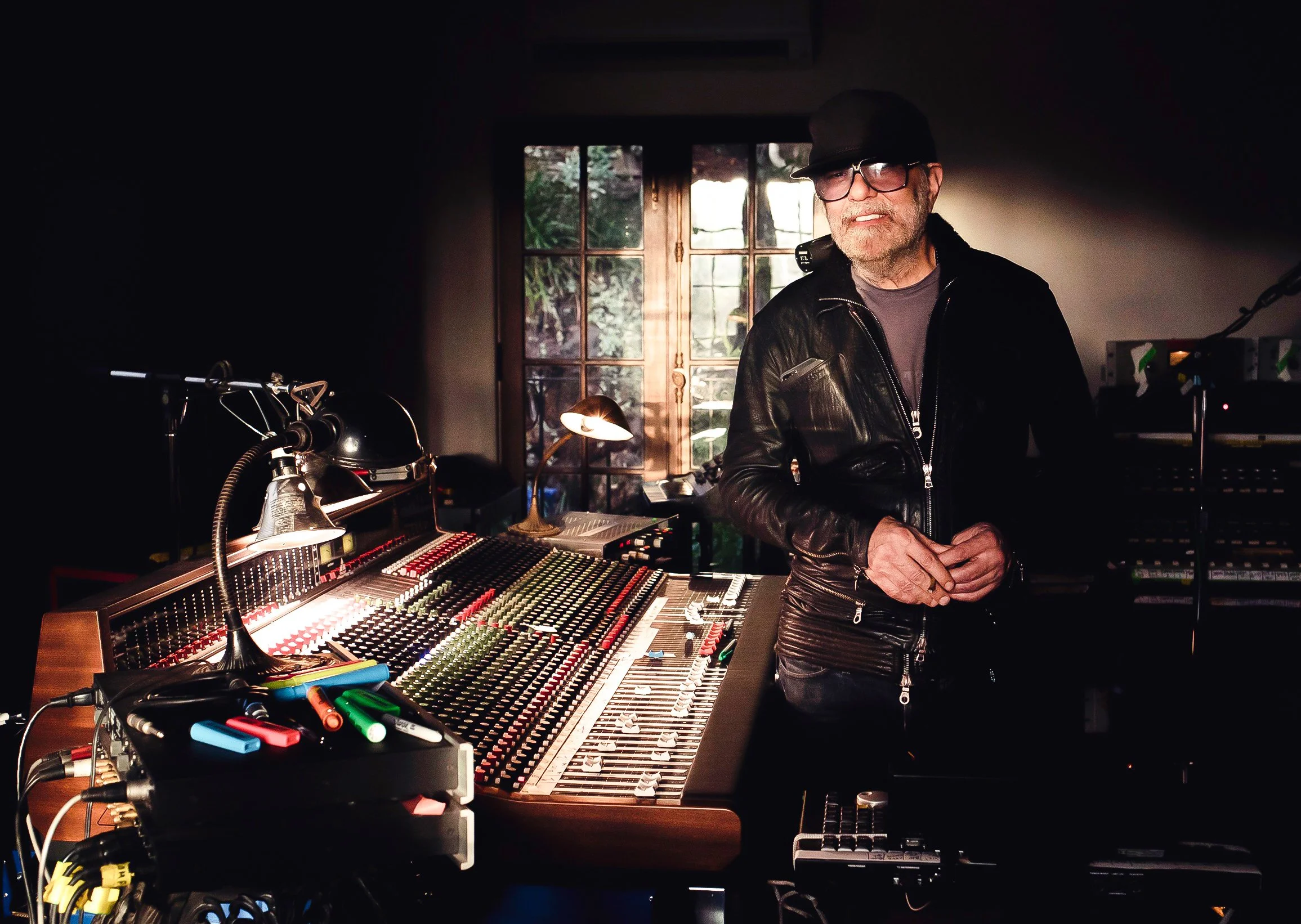 A man wearing glasses, a black cap, black leather jacket, and a gray shirt stands in a dimly lit music studio next to a large mixing console with colorful controls and sliders, with a window showing greenery outside in the background.
