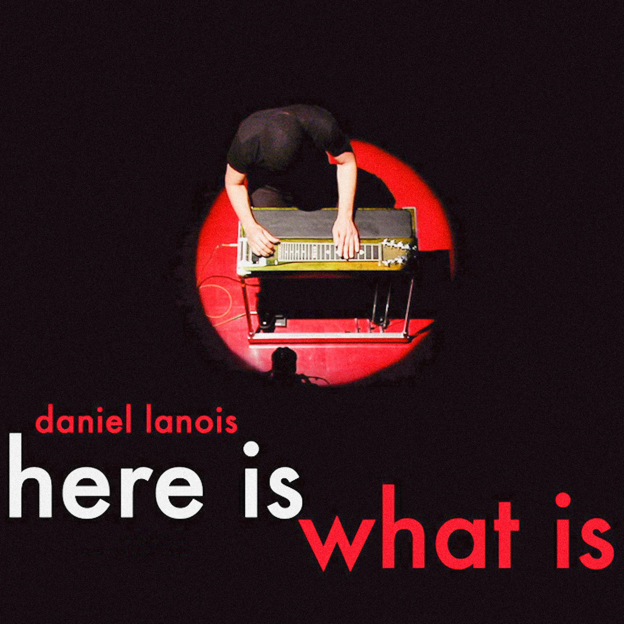 Top-down view of a musician playing a keyboard on a red stage, illuminated by a spotlight, with the text 'daniel lanois here is what is' in white and red.