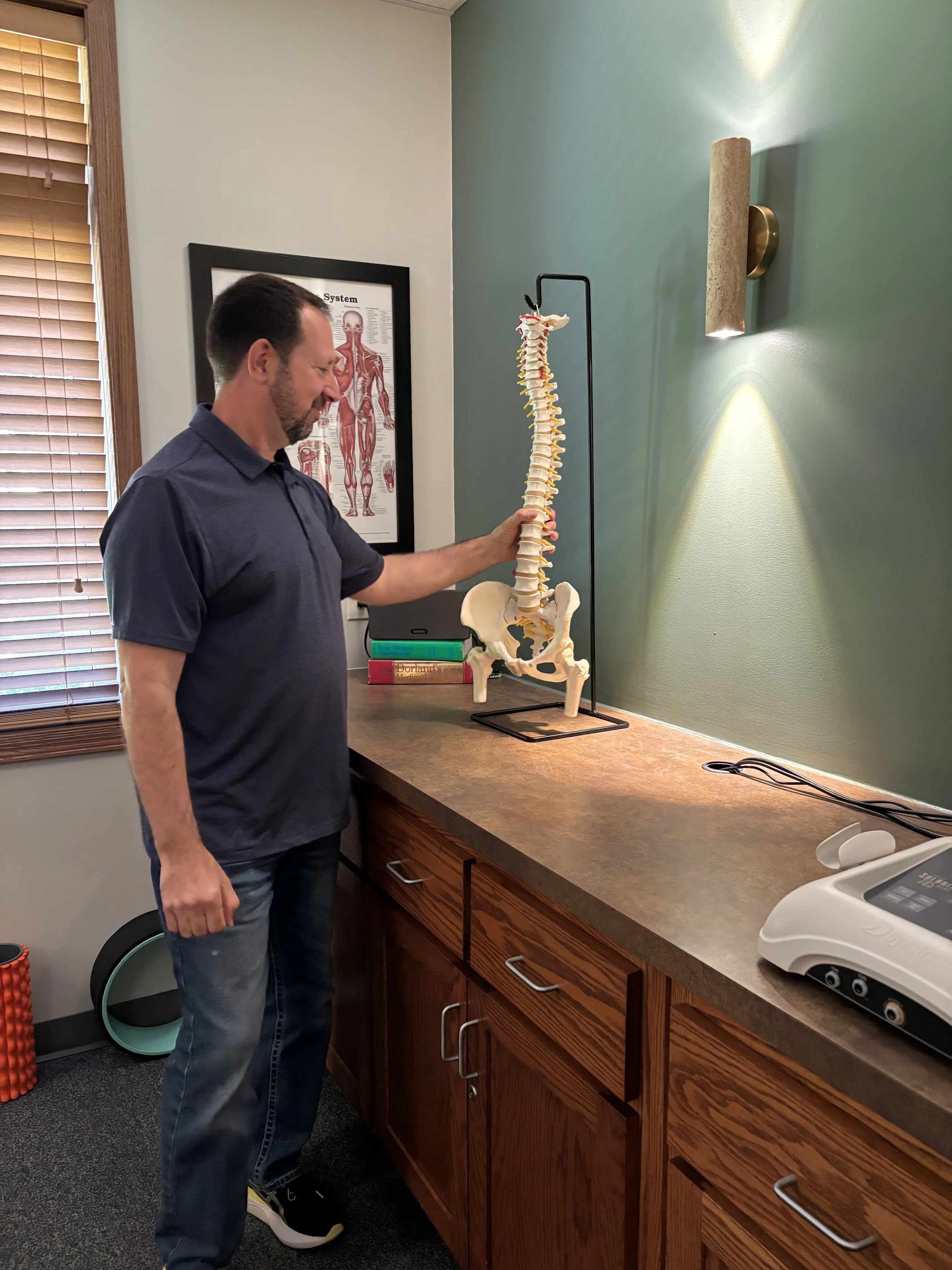 Dr. Pete Landry holding a spine model in a chiropractic treatment room.