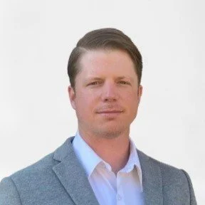 A man with short brown hair wearing a light gray blazer and a white shirt, standing against a plain white background.