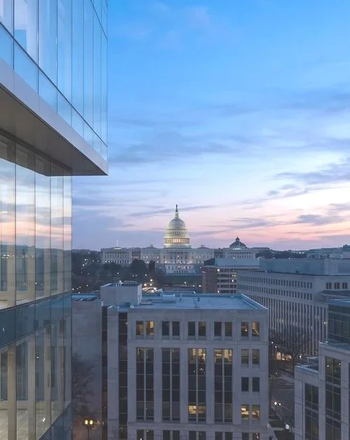 City skyline featuring the U.S. Capitol building under a partly cloudy sky during sunset, seen from a high-rise building with reflective glass windows.