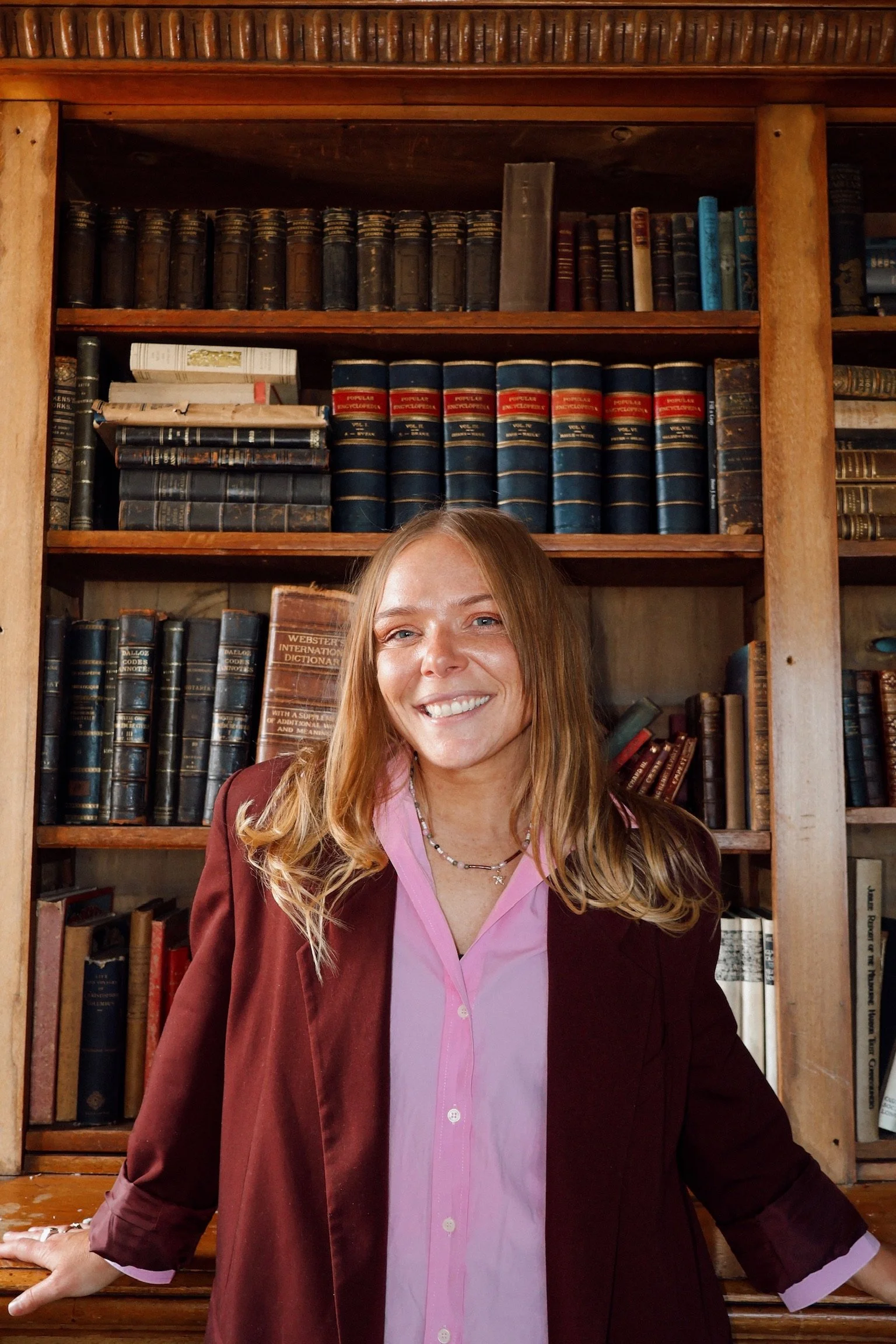 A woman with long, wavy red hair, wearing a maroon blazer over a pink button-up shirt, smiling at the camera, standing in front of a wooden bookshelf filled with old books.