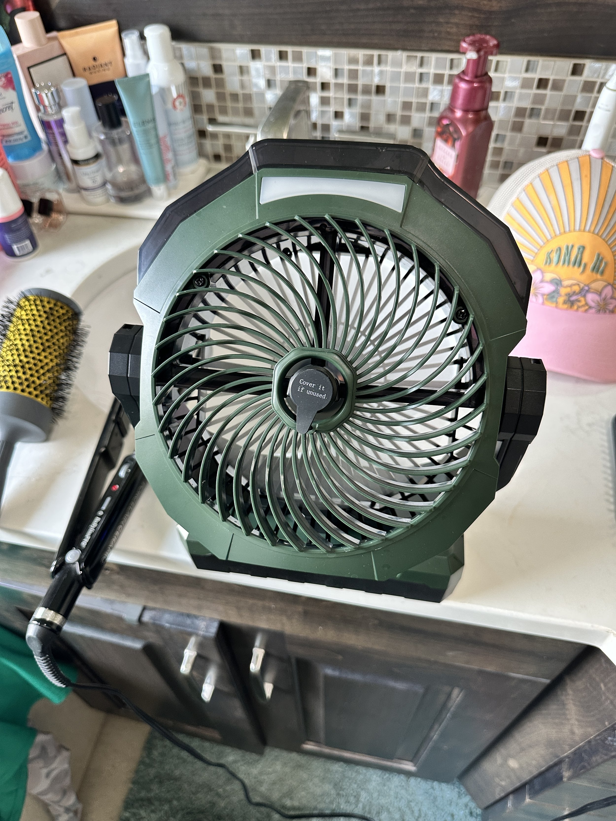 Close-up of a portable green and black fan sitting on a kitchen countertop in front of a soap dispenser and various bottles.
