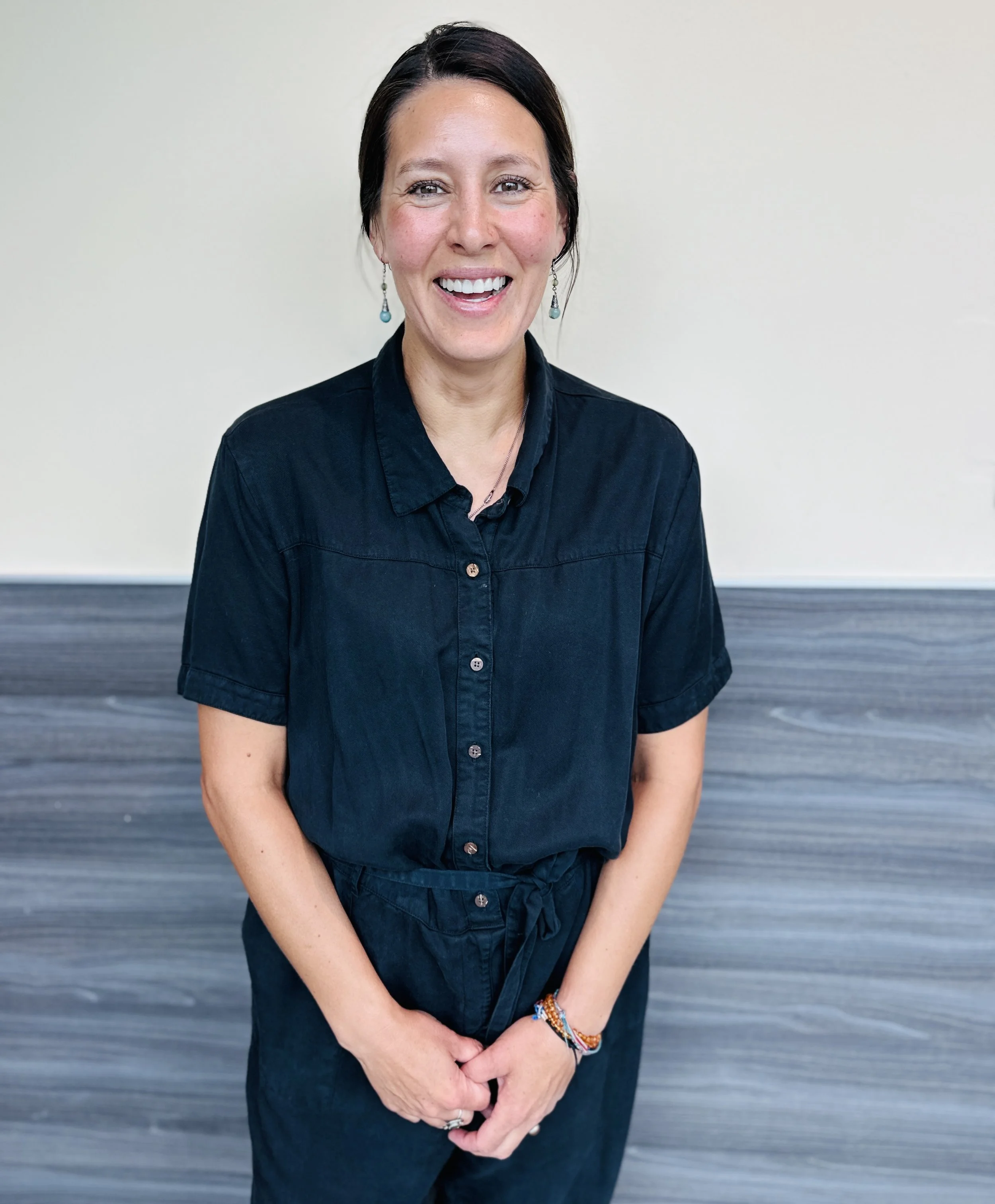 A woman with short dark hair and earrings smiling, wearing a black button-up shirt and dark pants, standing against a light-colored wall.