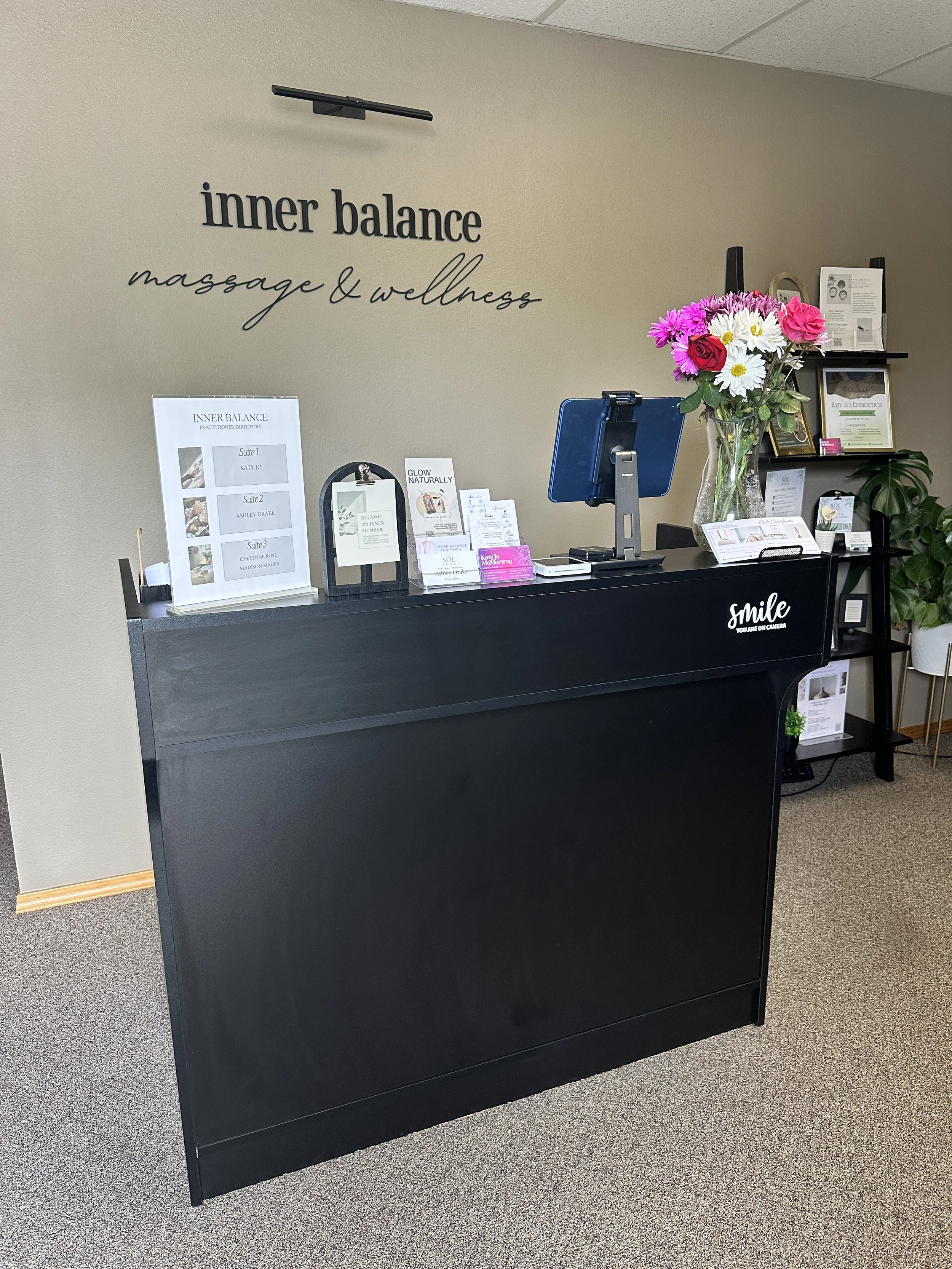 Reception desk with a large floral arrangement, informational flyers, and a computer monitor, inside a wellness center with beige walls and gray carpet.