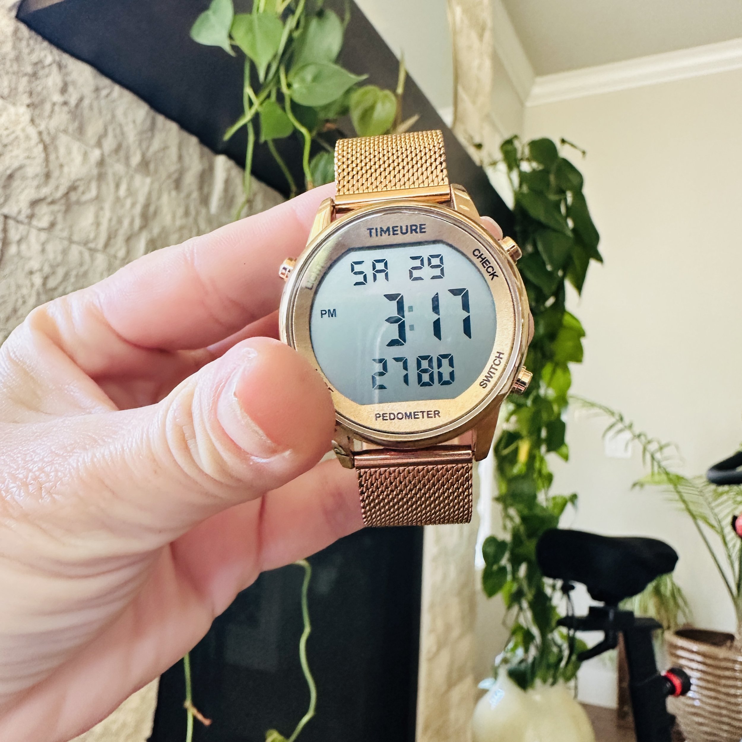 Close-up of a gold digital wristwatch with a mesh band displaying the date and time, held by a person's hand against a background of green houseplants and a black surface.