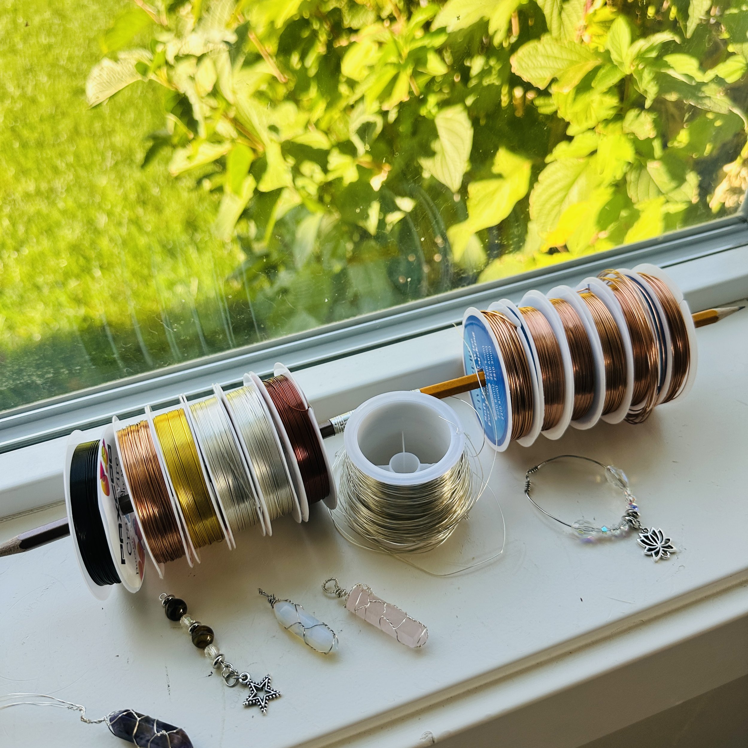 A window sill holding spools of copper wire and jewelry-making supplies, with greenery outside.