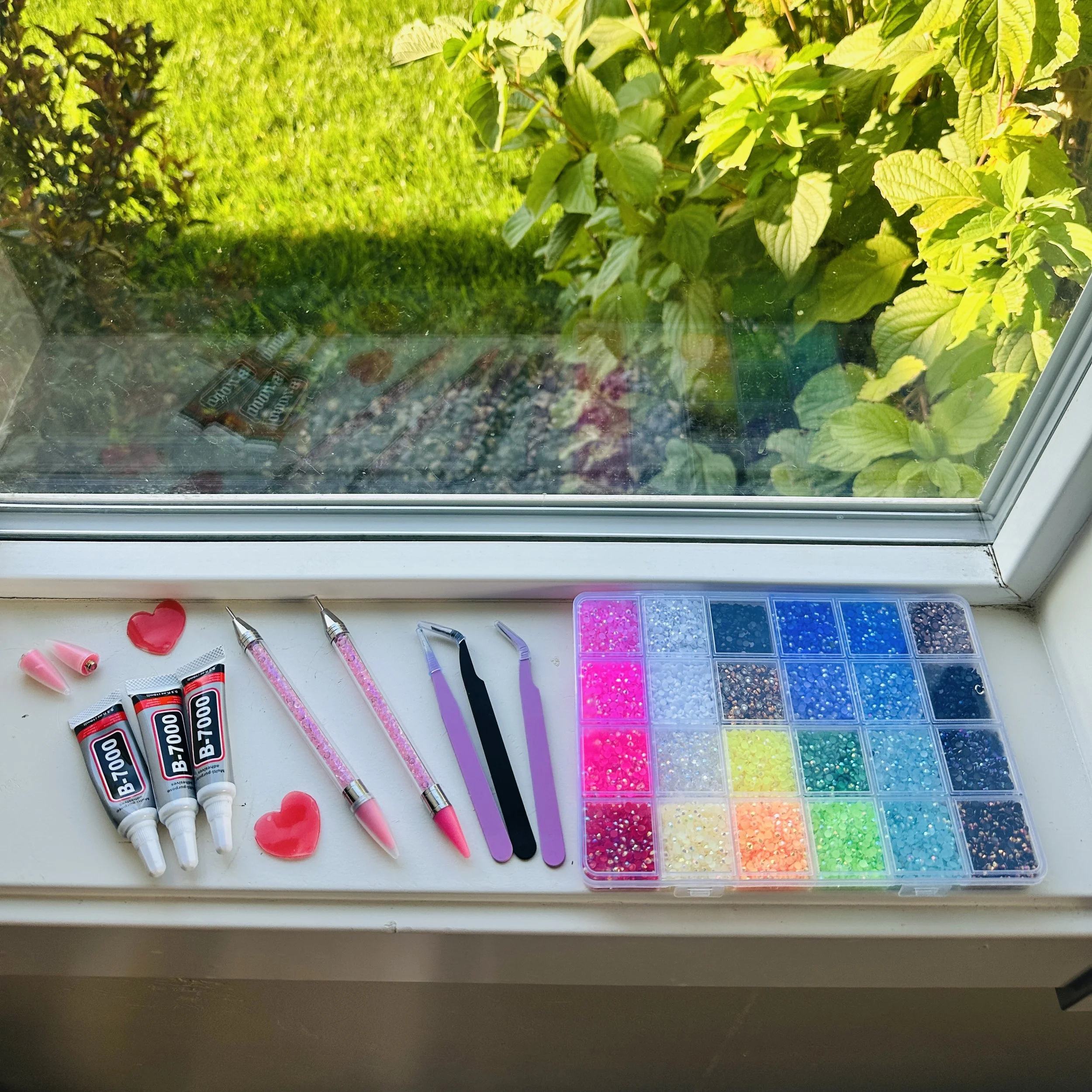 Assorted crafting supplies on a white table inside a room, including glue sticks, craft tools, and small plastic containers of colorful rhinestones.