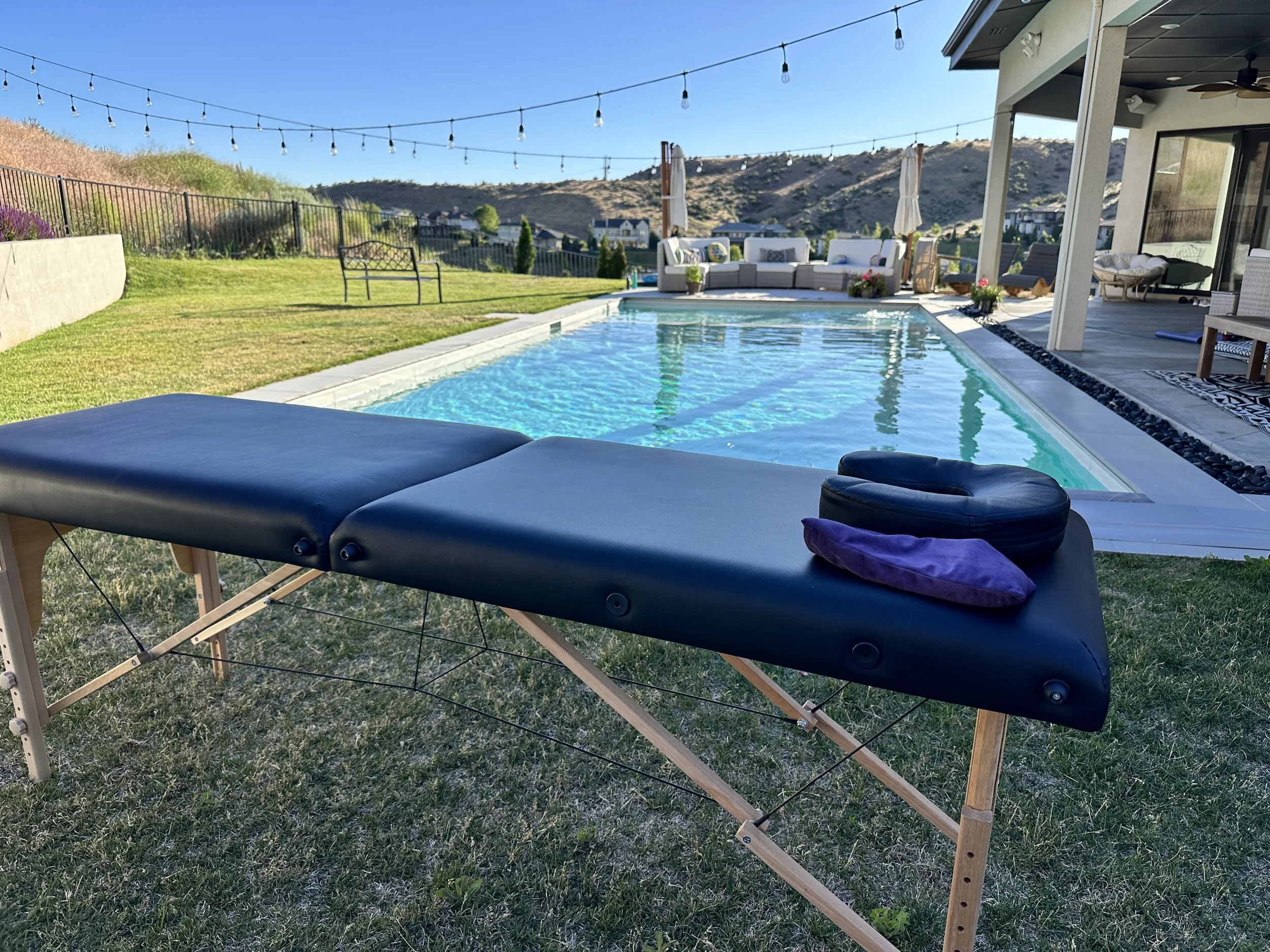 A backyard with a swimming pool, surrounded by a grassy lawn, outdoor seating with a sectional sofa and umbrellas, string lights overhead, and a mountain range in the background.