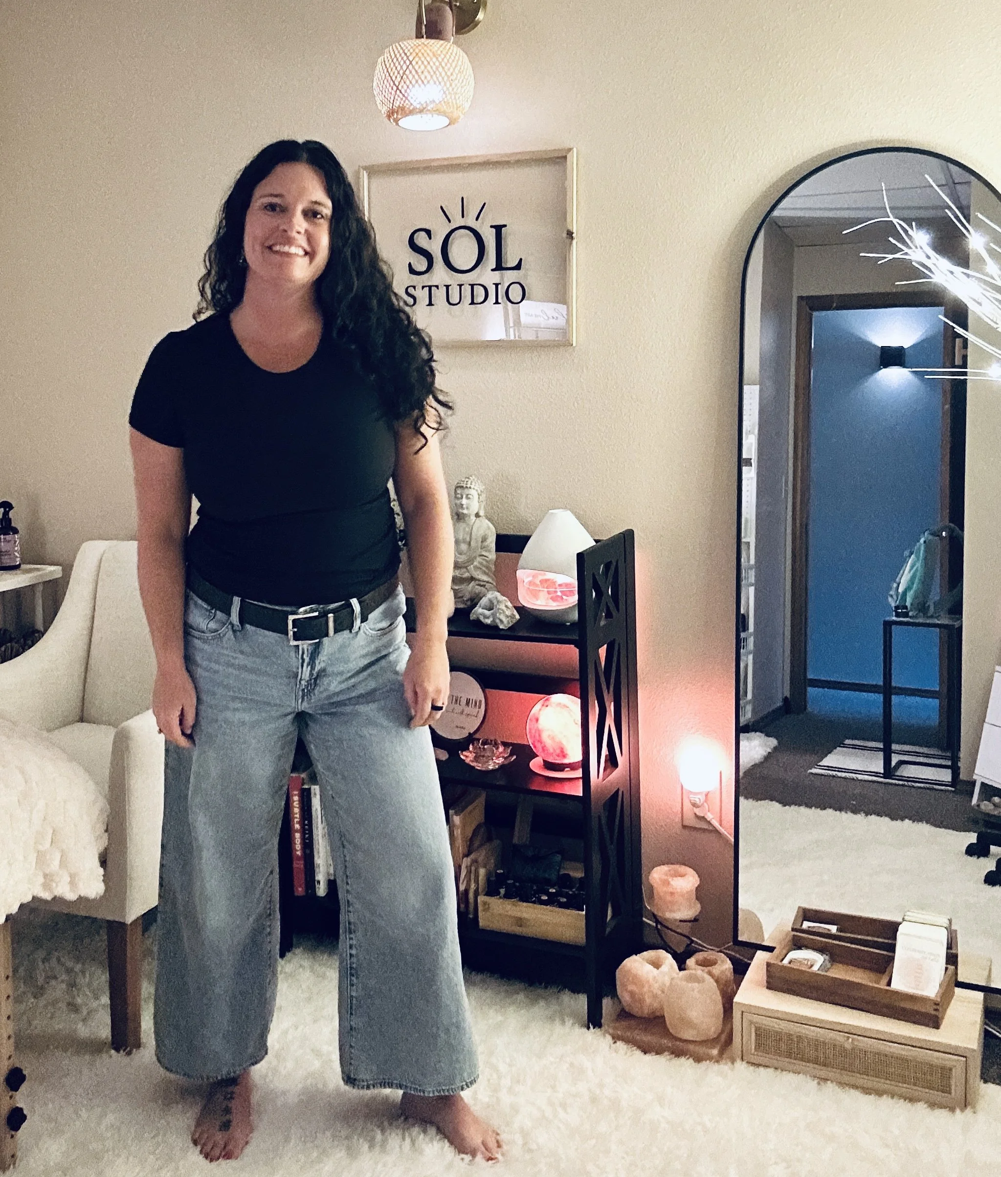 A woman standing in a cozy living room with a spiritual and wellness decor. She has long, curly dark hair, is wearing a black t-shirt and light blue jeans, and is barefoot on a white shaggy rug. Behind her, there's a white sofa, a bookshelf filled with books, and a Buddha statue. To the right, a black shelving unit displays Himalayan salt lamps, candles, and decorative rocks, with a lamp on top. The wall features a framed print with the words "SÓL STUDIO" and a simple sun illustration. There is a mirror reflecting part of the room, including a doorway with a blue wall.