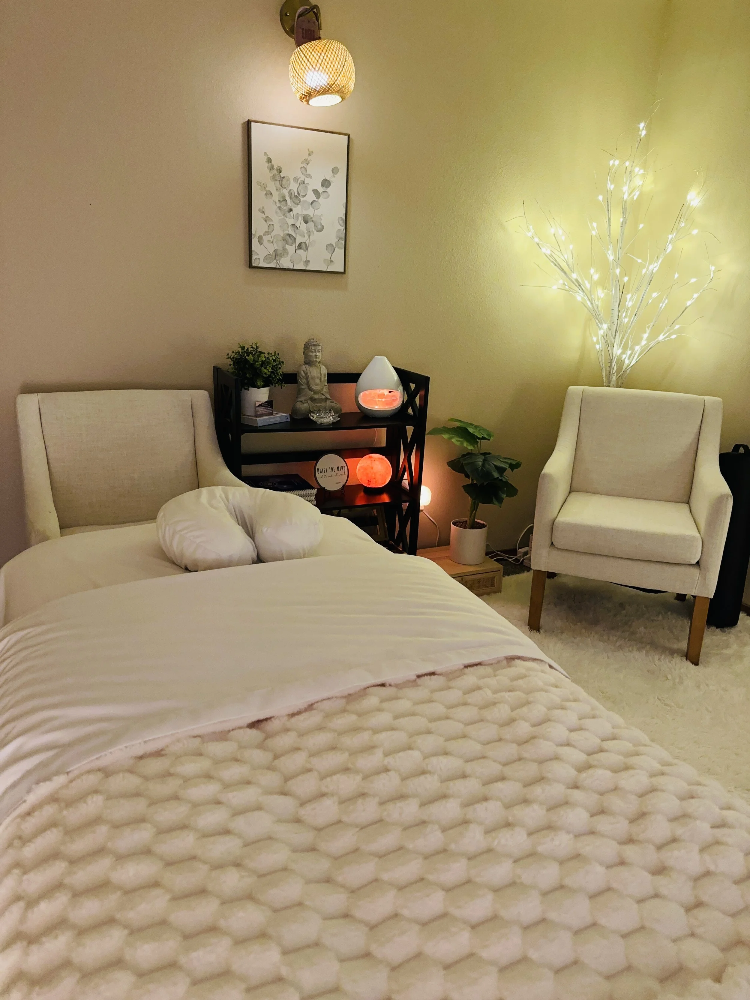 Cozy bedroom with a white bed, a beige armchair, a small black shelf with decorative items, potted plants, and ambient lighting including a glowing white tree and a Himalayan salt lamp, with a framed botanical print on the wall.