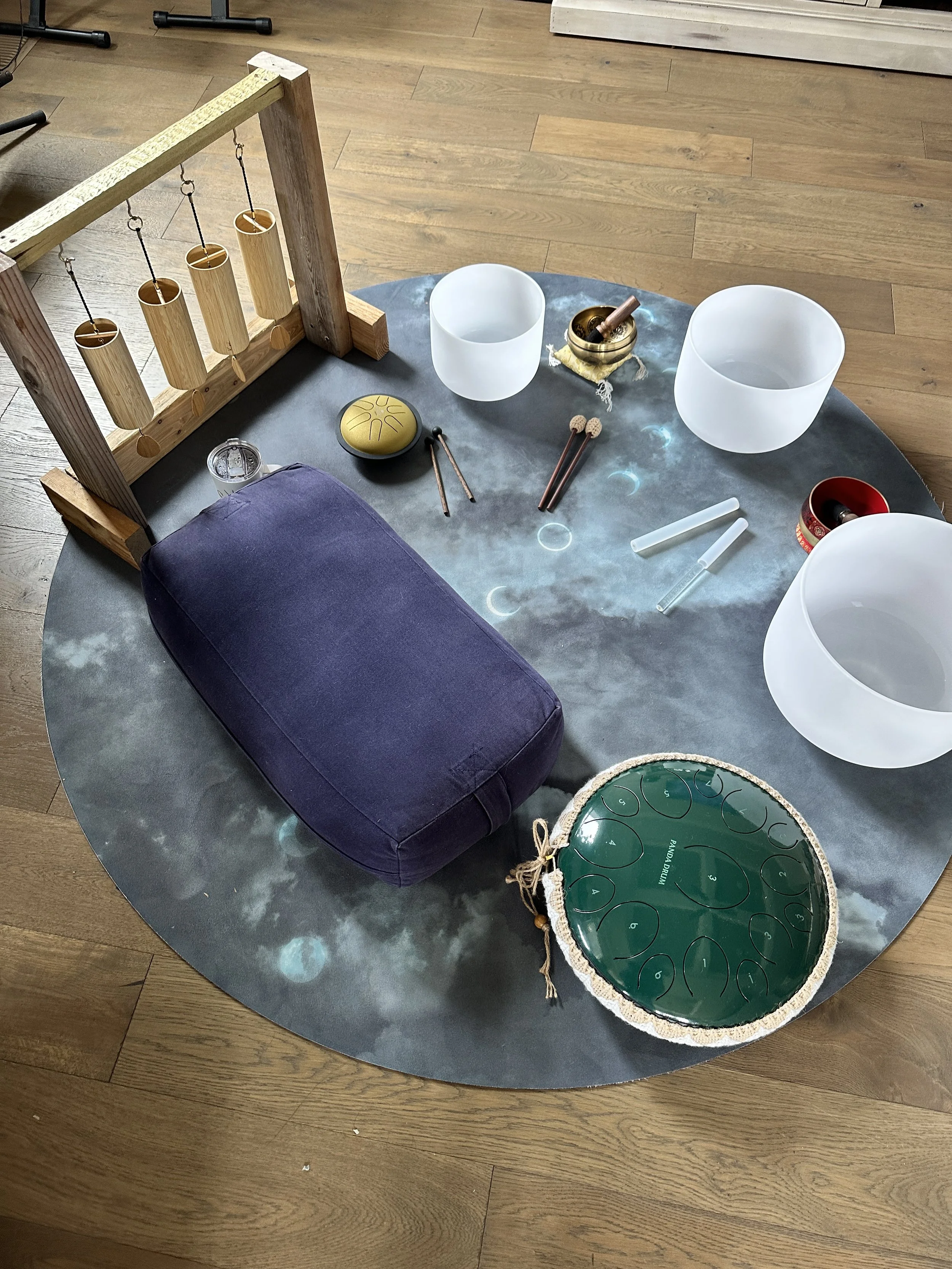 A round table with singing bowls, a purple cushion, a wooden frame with hanging chimes, and a moon phase chart on a wooden floor.
