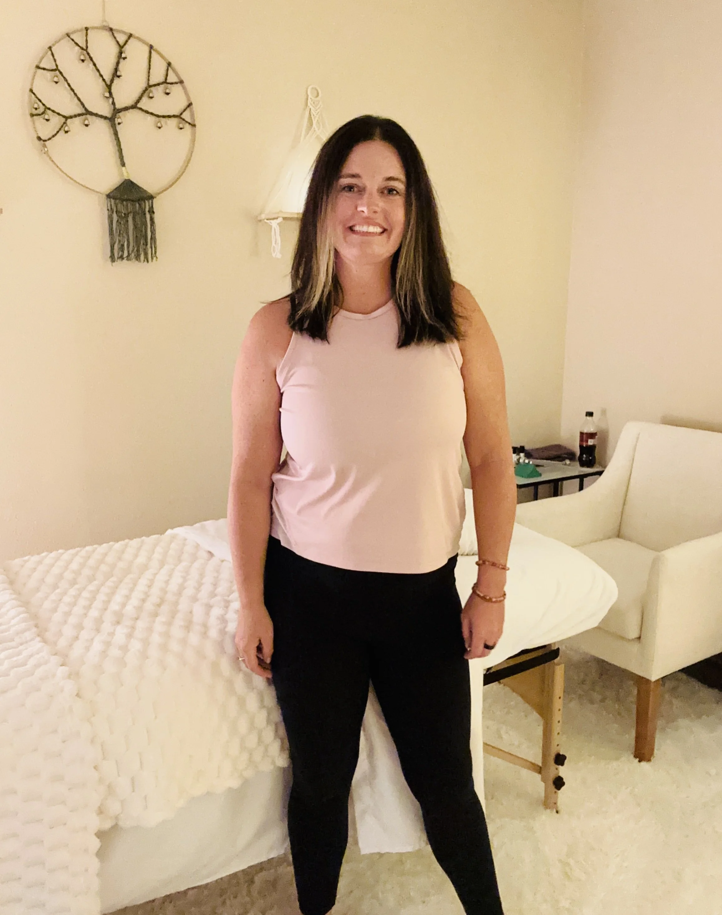 A woman with long dark hair and a light pink sleeveless top standing in a bedroom in front of a white bed with a textured comforter. There is a wall decor piece like a circular metal tree on the wall to her left, a wall lamp behind her, and a table with drinks and items to her right.