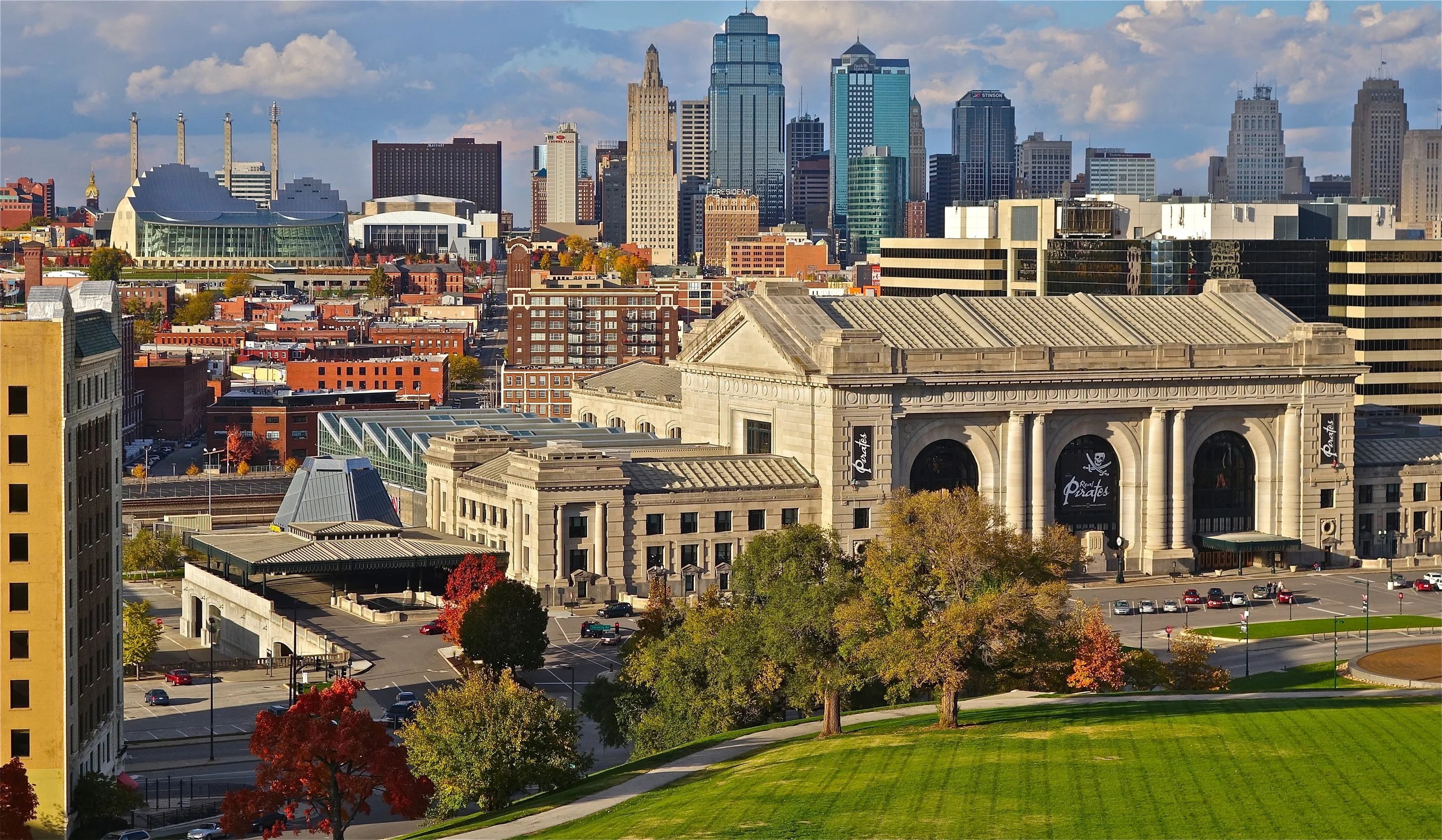 City skyline with tall skyscrapers, a historic building with columns, trees in fall colors, and park area in foreground.
