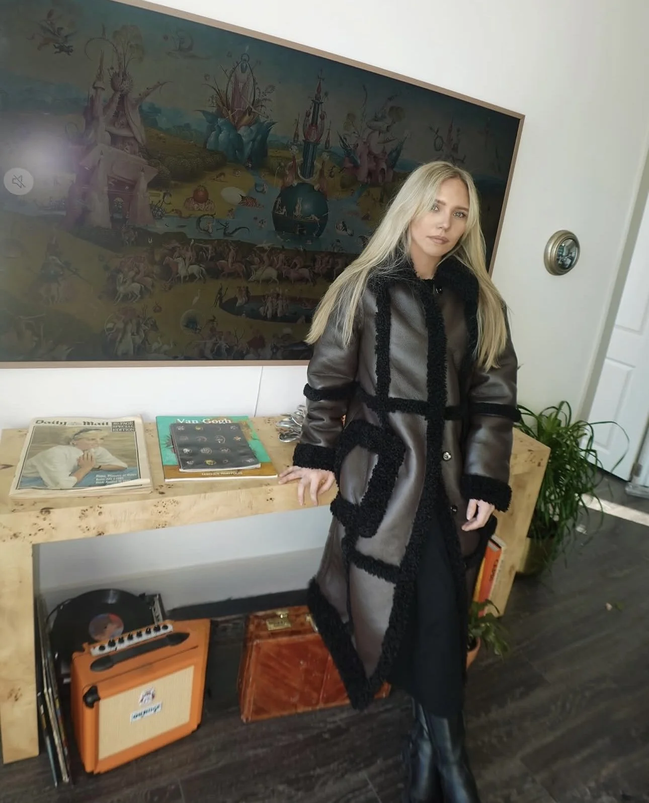 A woman with long blonde hair wearing a gray and black coat standing in front of a wooden table with magazines, an orange mini amplifier, and a small wooden box. Behind her is a wall with a painting and a potted plant, and on the table are also vinyl records and a turntable.