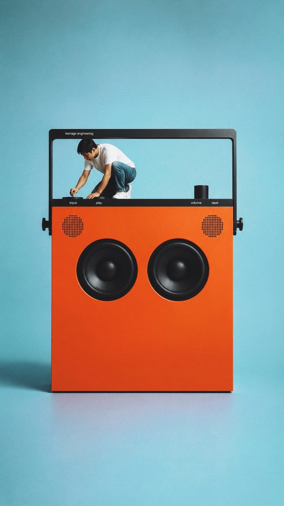 A young boy is on top of a large speaker that is designed to look like a vintage boombox, with a bright orange front and large black speakers. The speaker is set against a light blue background.