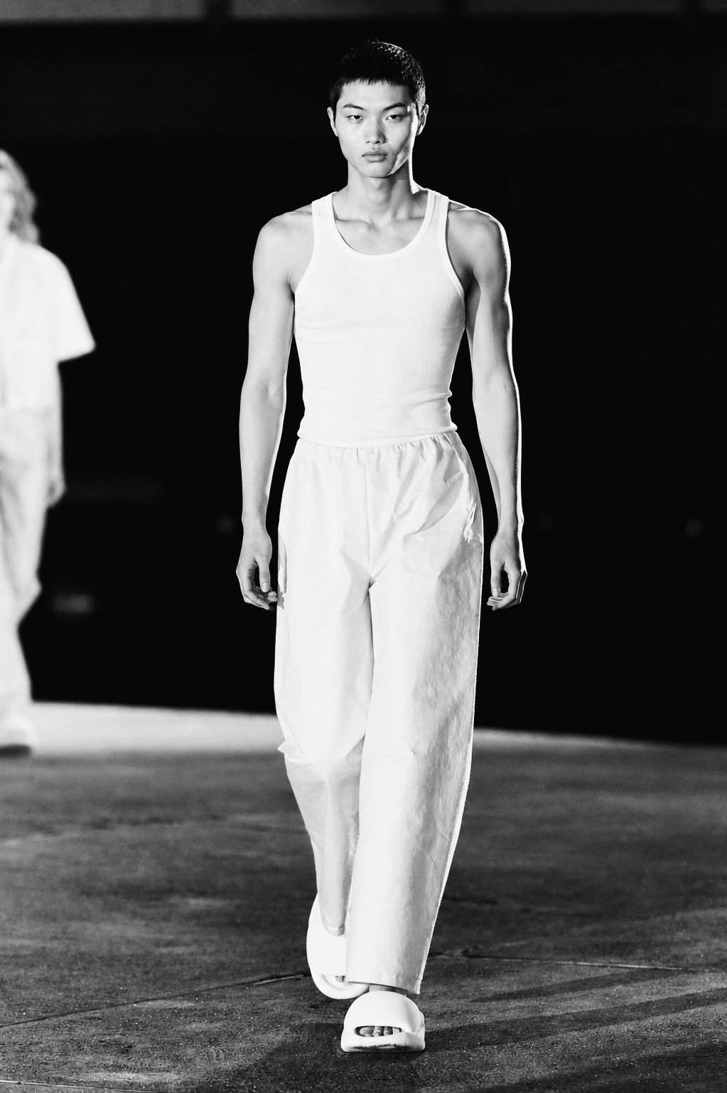 A black-and-white photo of a young Asian man walking on a runway, wearing a white tank top, loose white pants, and platform sandals, with a serious expression.
