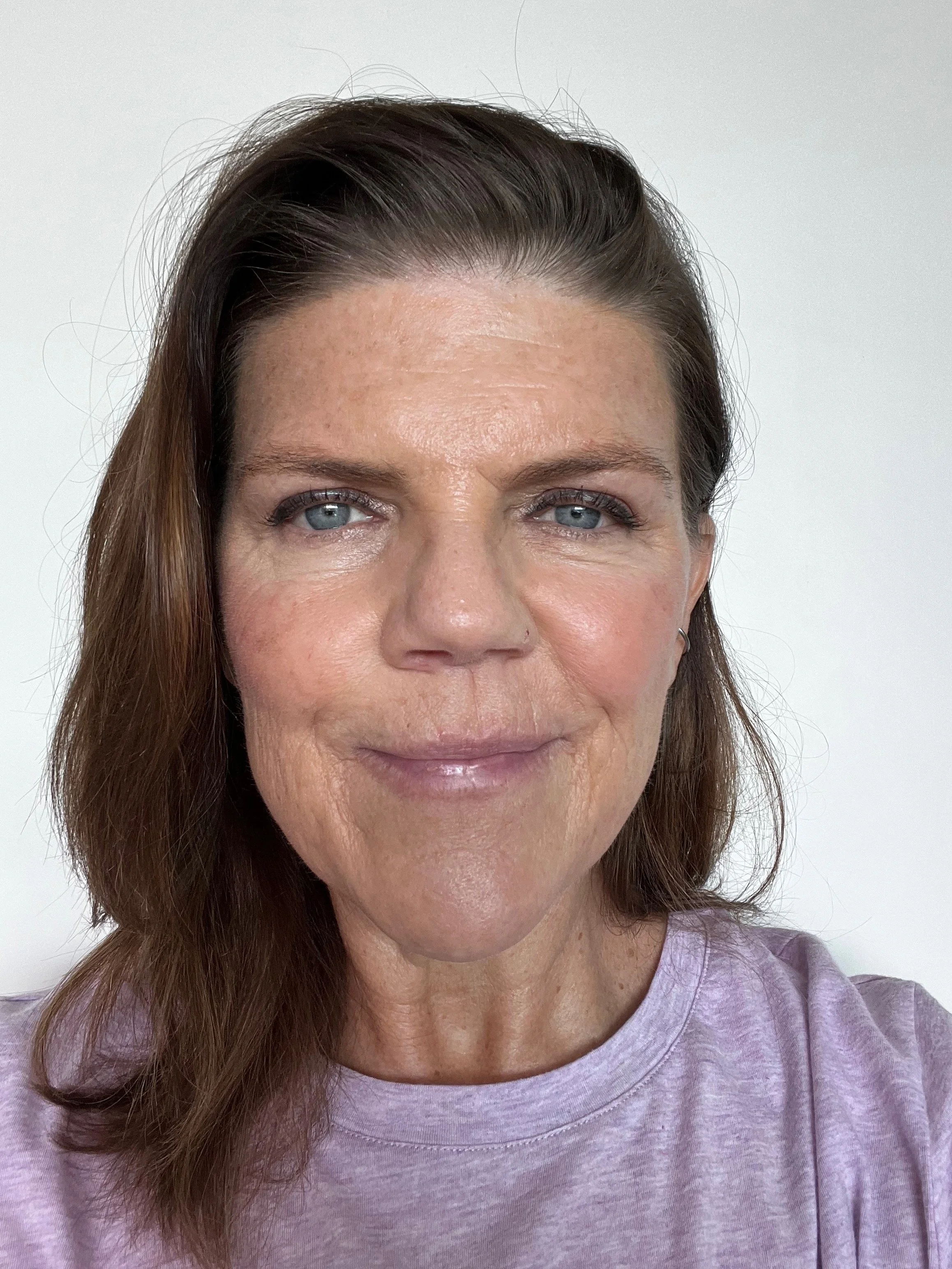 Close-up of a woman with long brown hair and light blue eyes, wearing a light purple shirt, smiling against a plain white background.