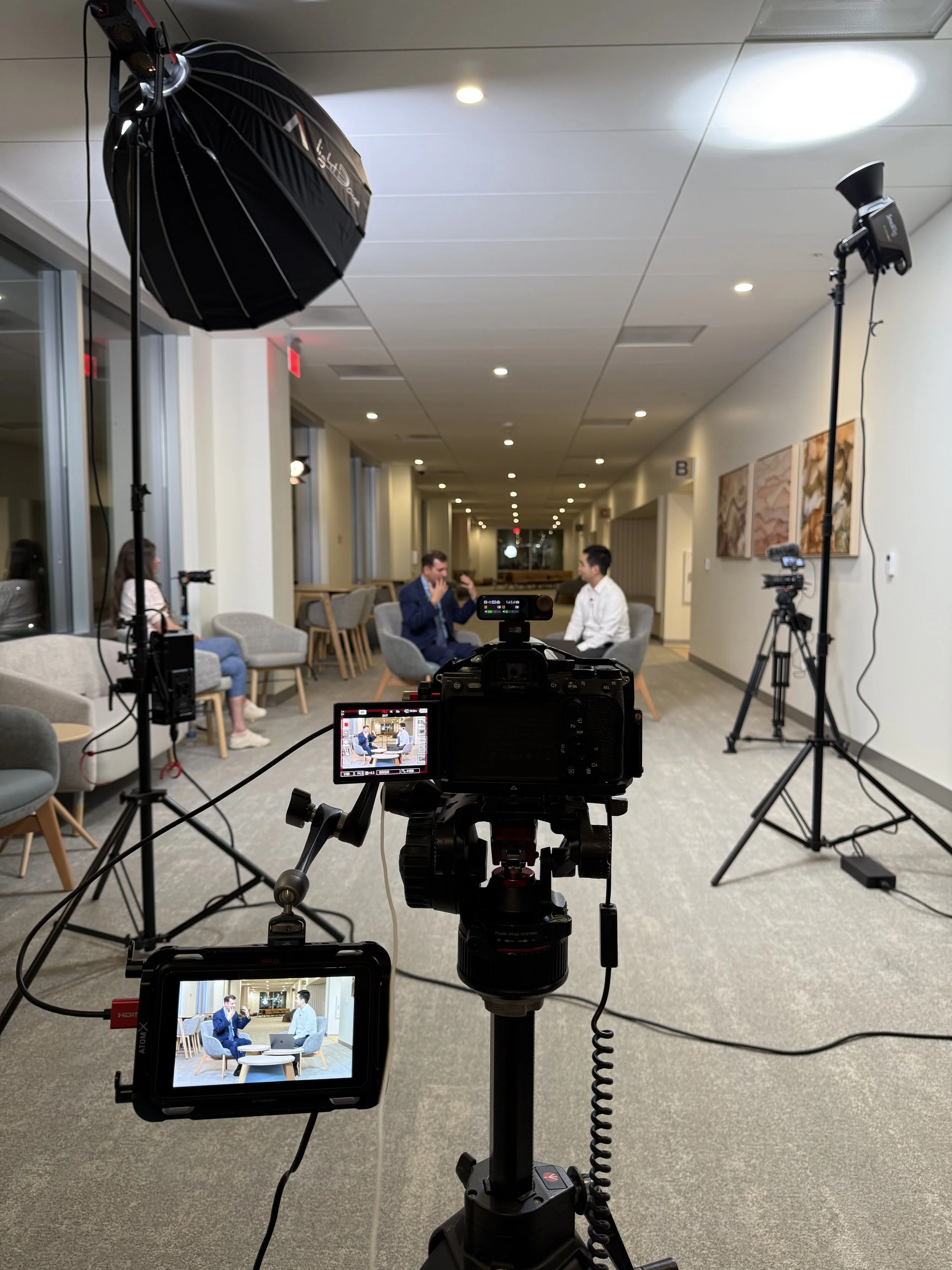 Studio setup with a professional camera on a tripod recording two men having a conversation in a modern, well-lit lounge area with artwork on the wall and several chairs and tables.