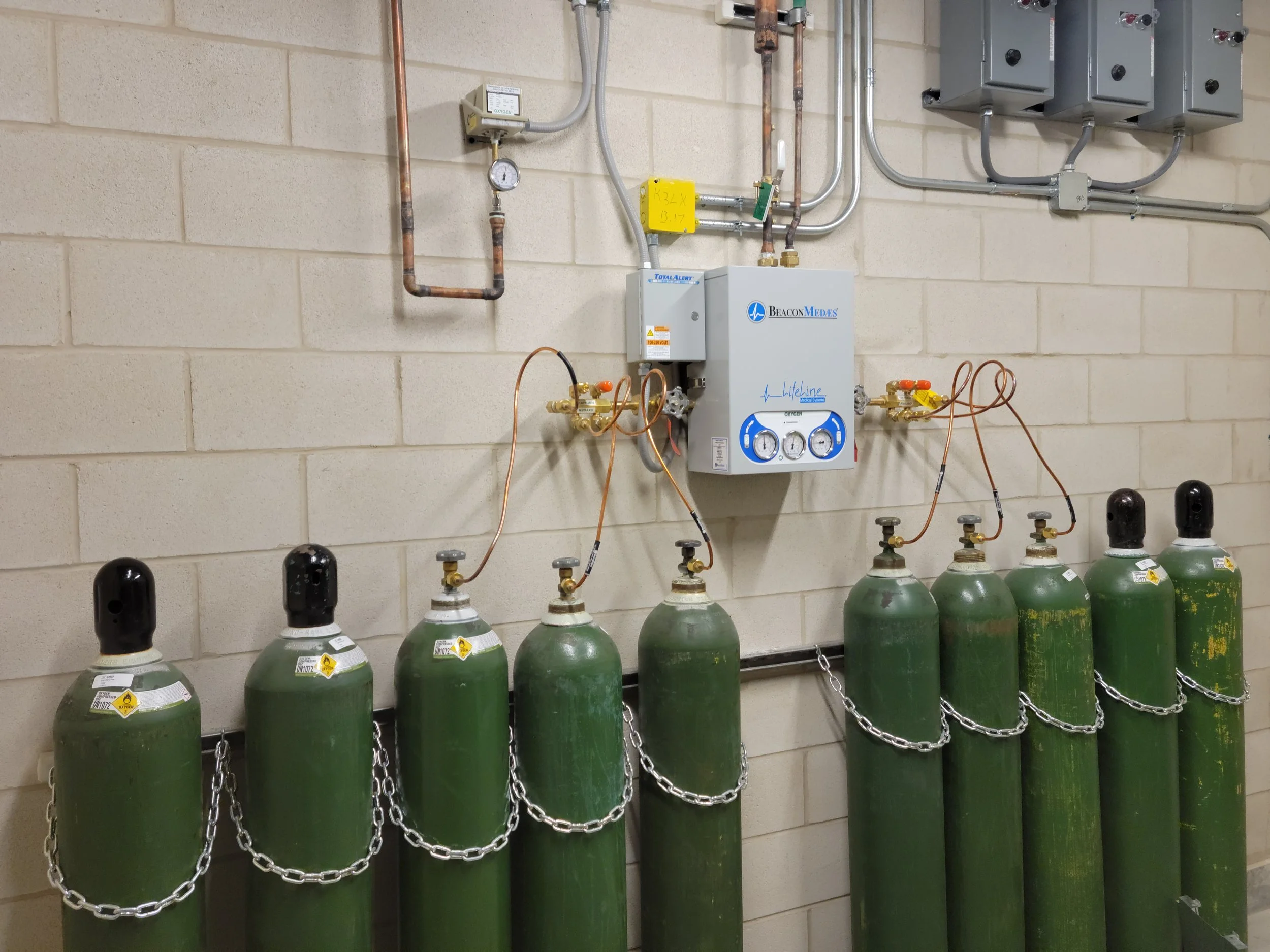 Medical oxygen tanks lined up against a beige brick wall, connected to a central oxygen regulator system with gauges, valves, and tubing.