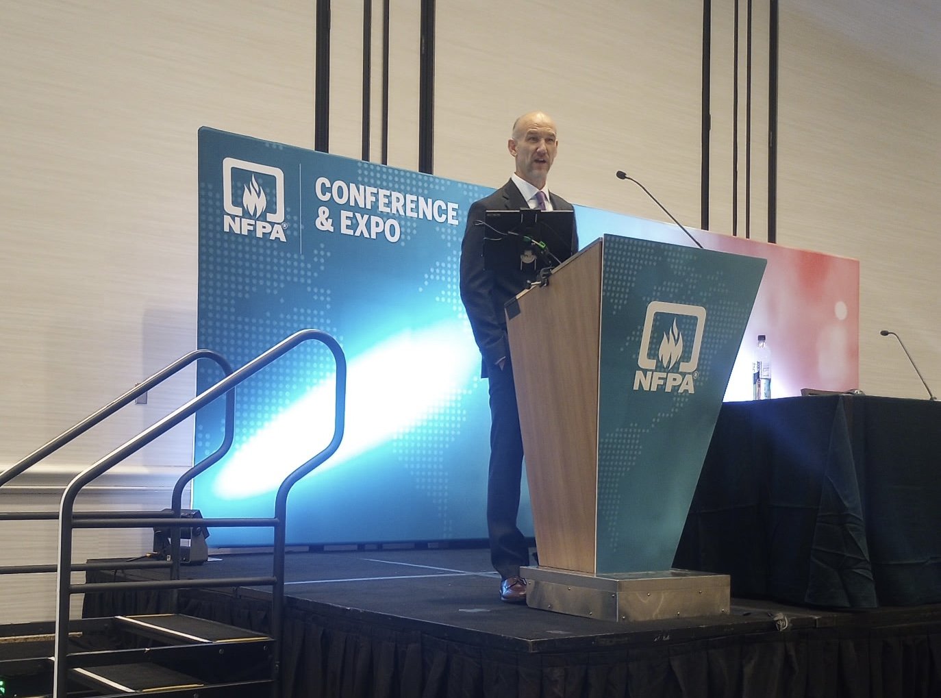 A man in a suit speaking at a podium during a conference and expo organized by NFPA, with NFPA banners behind him.