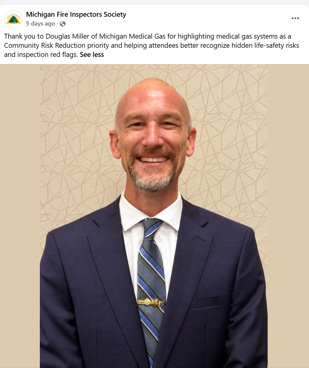 Smiling man in a navy suit, white shirt, and striped tie standing against a beige geometric patterned background.
